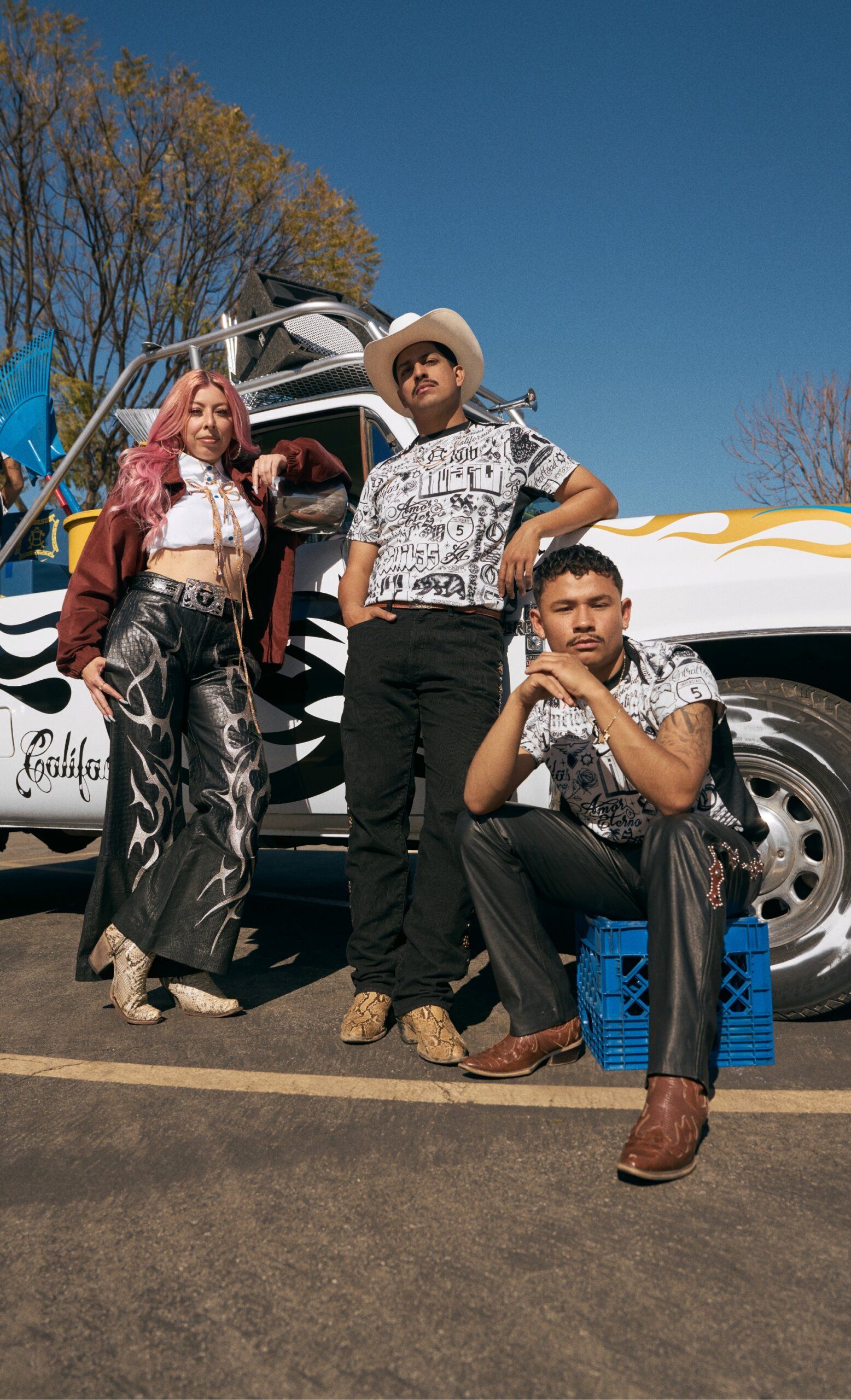 Three people pose in front of a white truck with custom black and yellow designs; one leans on the truck, one stands, and one crouches on a blue crate. It's a sunny day.