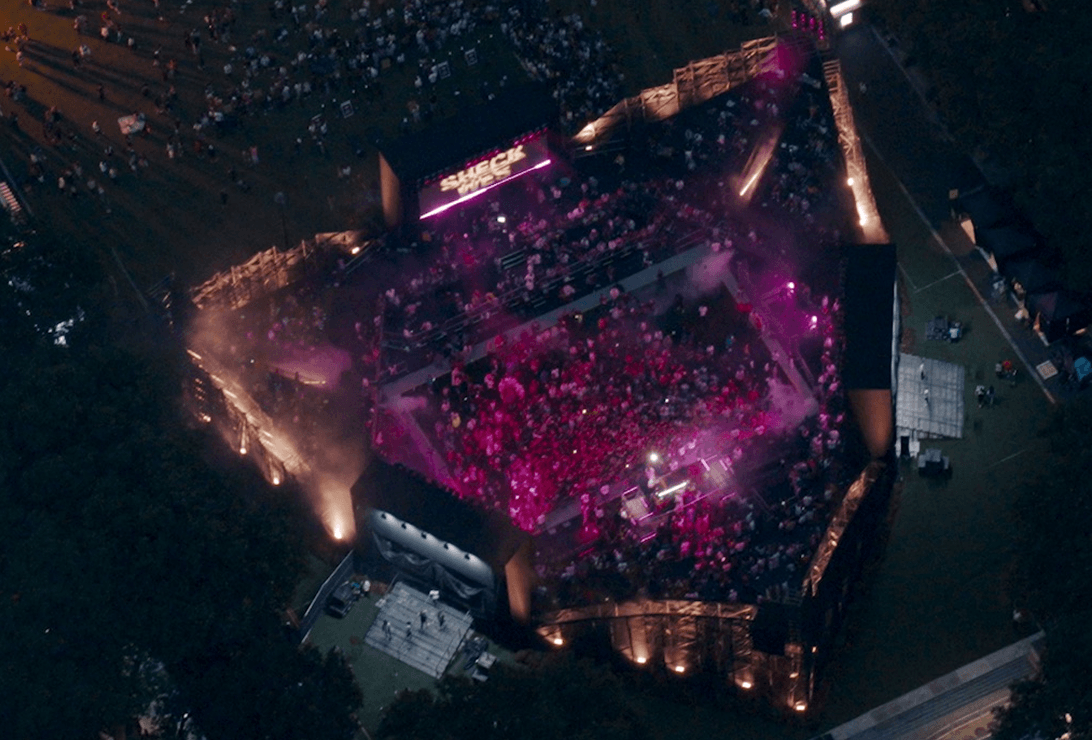 Aerial view of a large outdoor concert stage lit with purple lights, surrounded by a crowd of people at night, with trees and open grassy areas nearby.