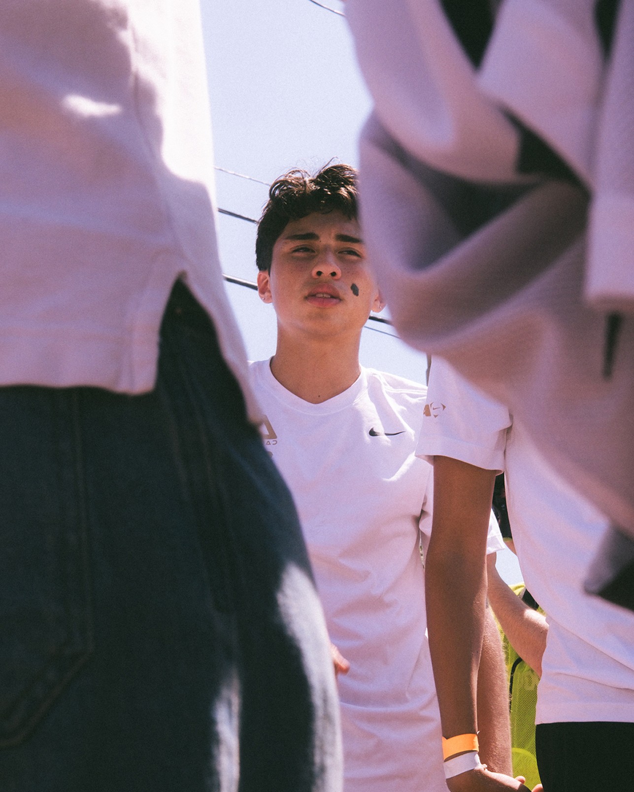 Teenage boy in a white shirt stands outdoors, partially framed by people in the foreground, with power lines and blue sky visible above.