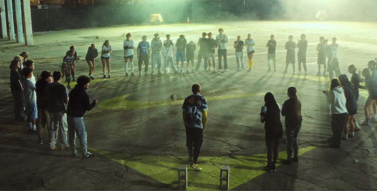 A group of people stands in a large circle outdoors at night, with one person in the center facing away from the camera. The scene is lit by diffuse, artificial light.