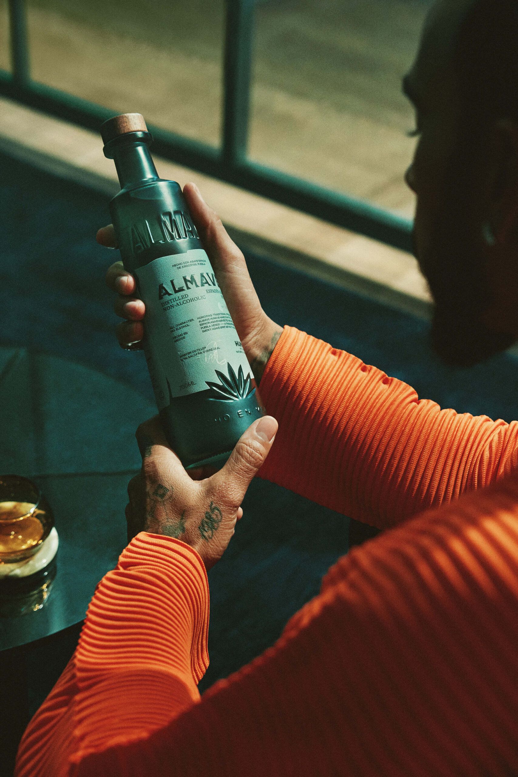 Person in an orange pleated shirt holds a green bottle labeled "Alma" with an agave illustration; a glass with a drink and ice sits nearby.