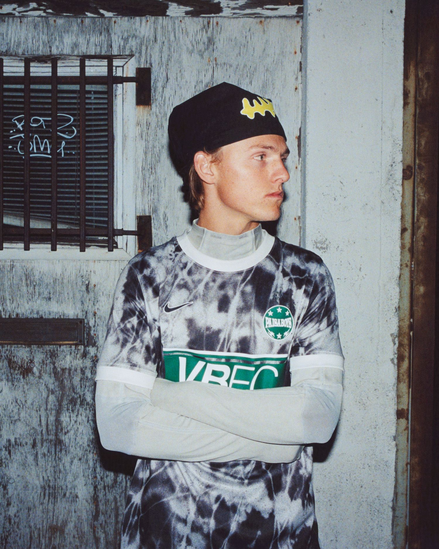 A person in a tie-dye football jersey and black cap stands with arms crossed in front of a weathered door, looking to the side.