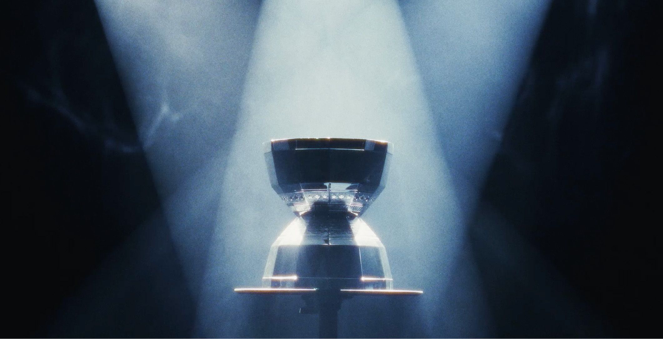 A silver trophy displayed on a pedestal is illuminated by intersecting spotlights in a dark, smoky environment.