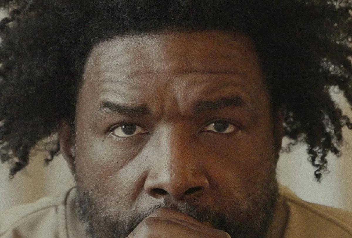 A man with curly hair and a beard looks directly at the camera, resting his chin on his hand.