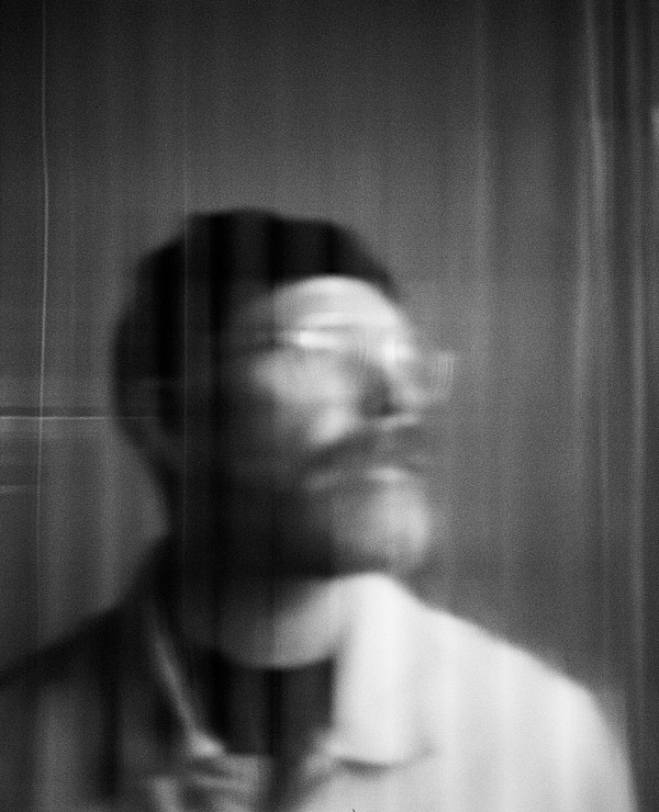 A blurry black and white photo of a man wearing glasses and a beanie, seen through a semi-transparent curtain with vertical lines.