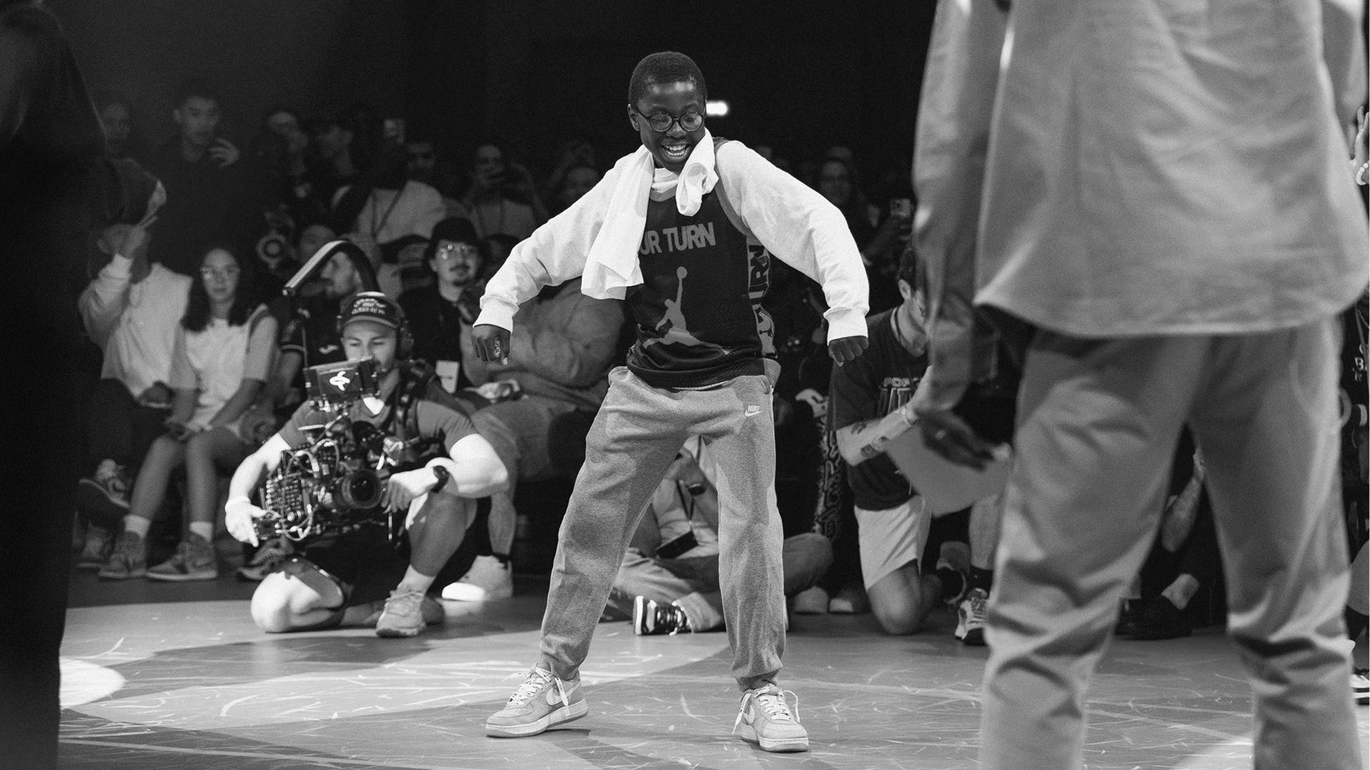 A person in casual clothes dances confidently in the center of a crowd, with photographers and spectators watching and capturing the moment.