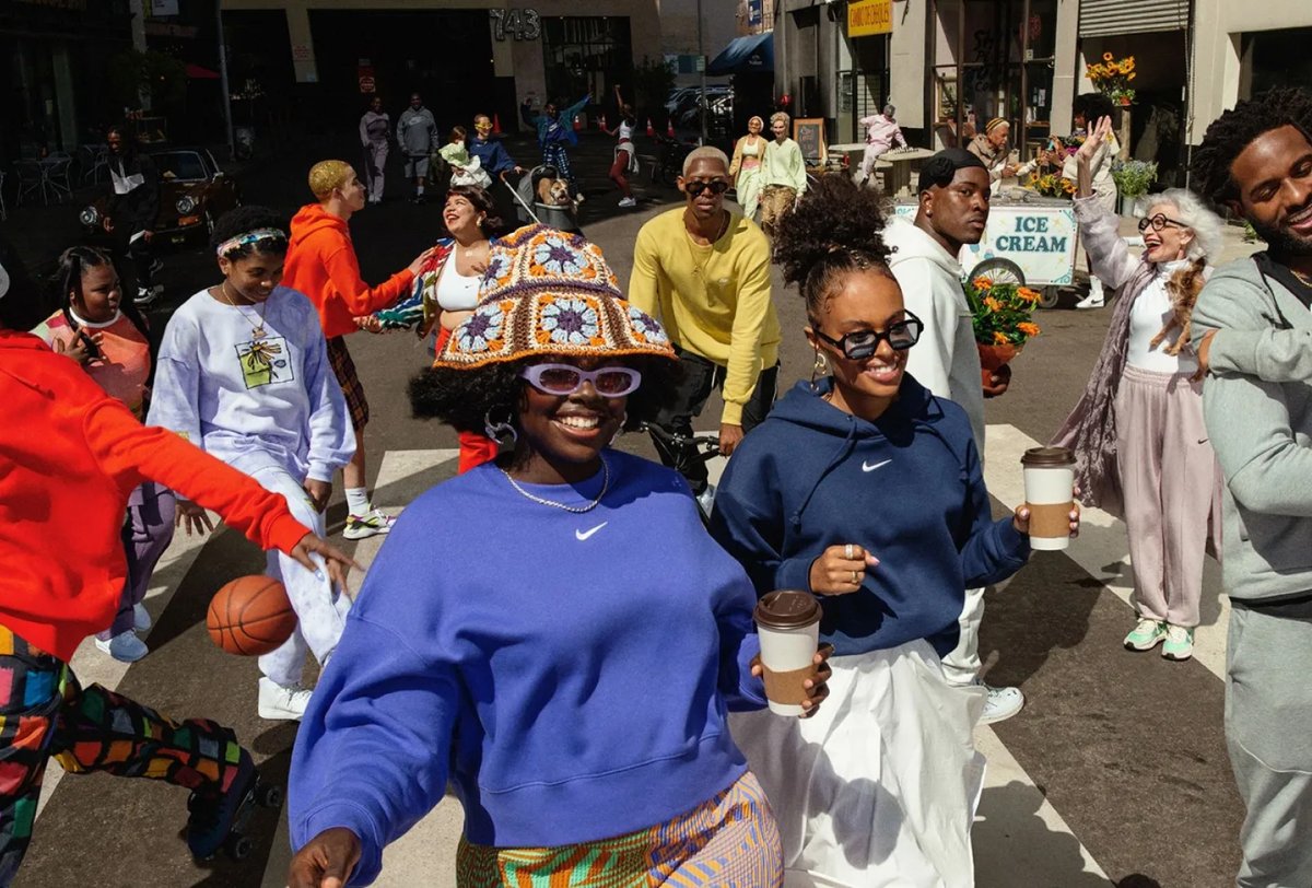 A diverse group of people cross a city street, some holding coffee or a basketball. Two women in the foreground smile and wear sunglasses and colorful outfits.