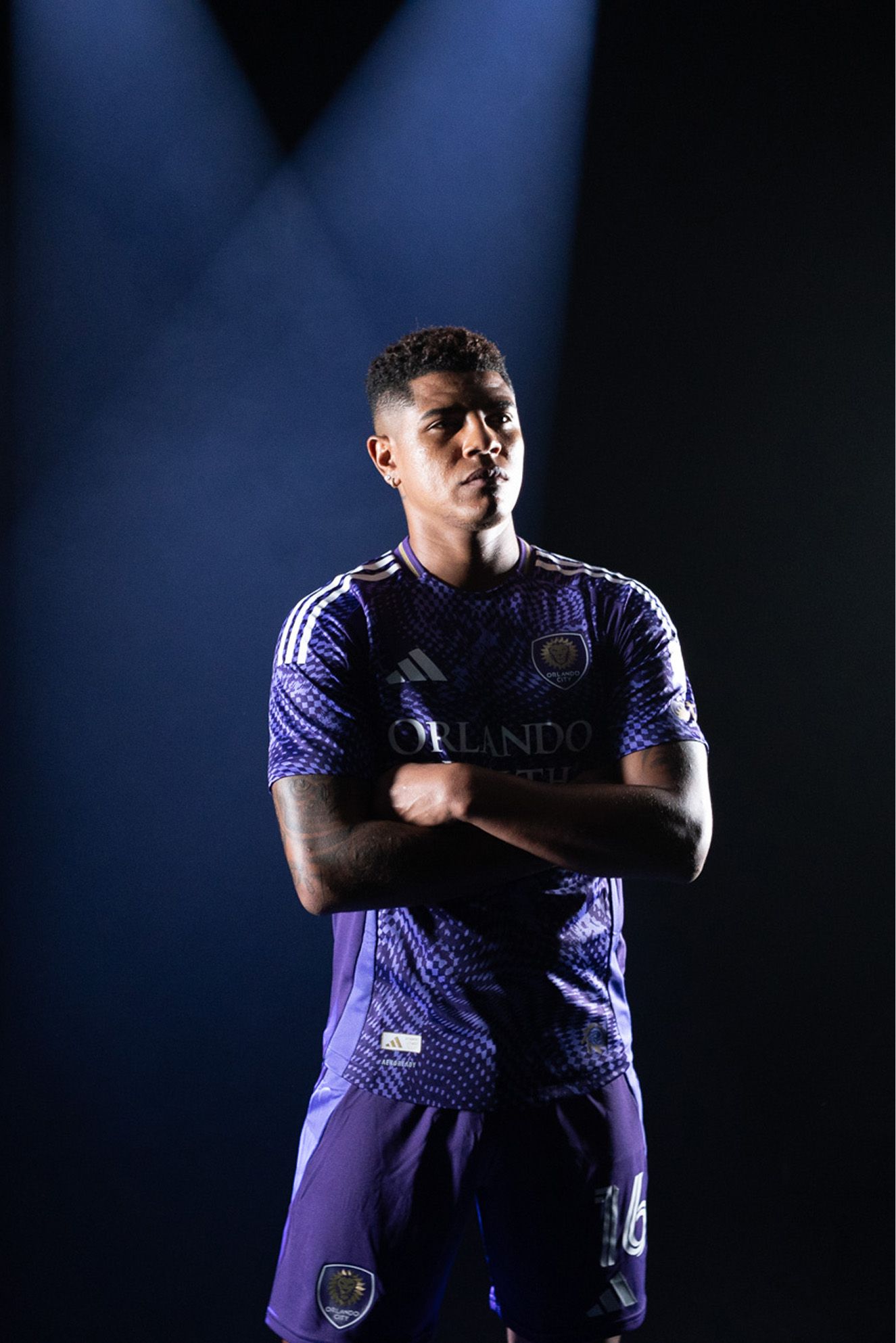 A soccer player in an Orlando City SC purple uniform stands with arms crossed under a spotlight against a dark background.