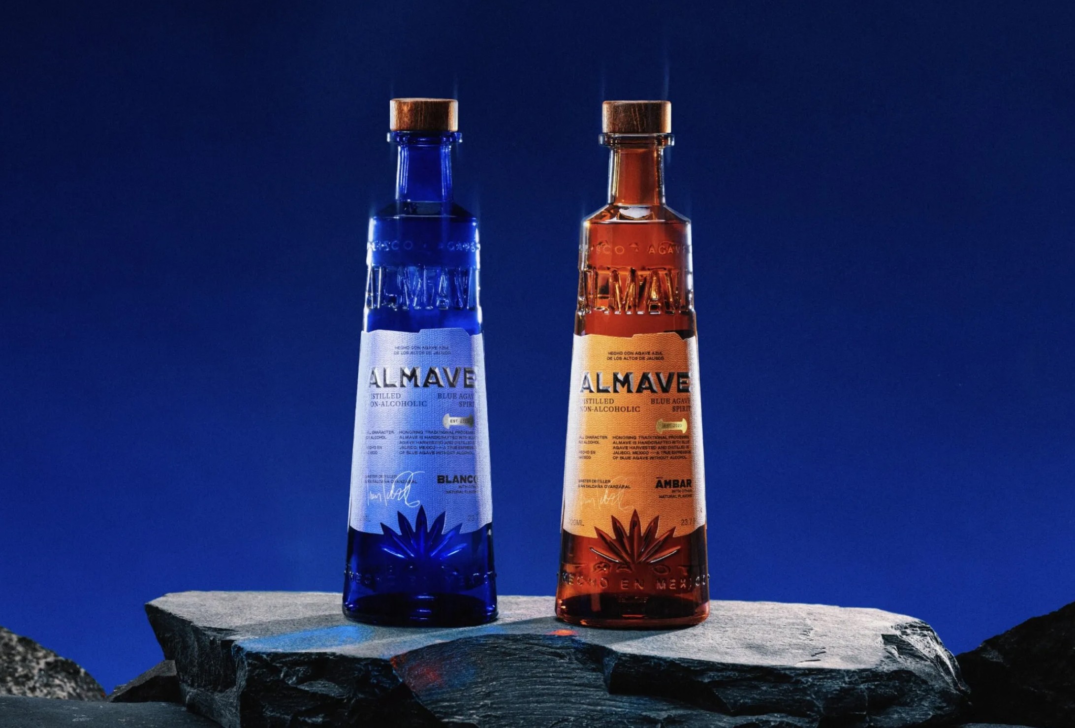 Two tall glass bottles, one blue and one amber, labeled "ALMAVÉ," stand side by side on flat rocks against a dark blue background.