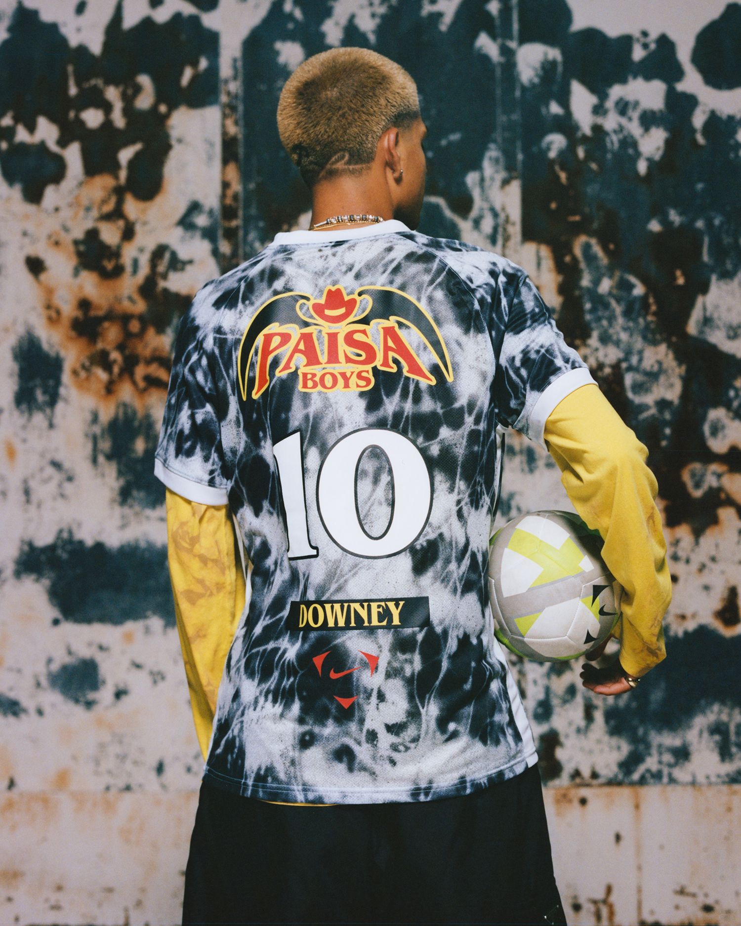 Person standing with their back facing the camera, wearing a black and white tie-dye “Paisa Boys” soccer jersey with the number 10 and holding a soccer ball.