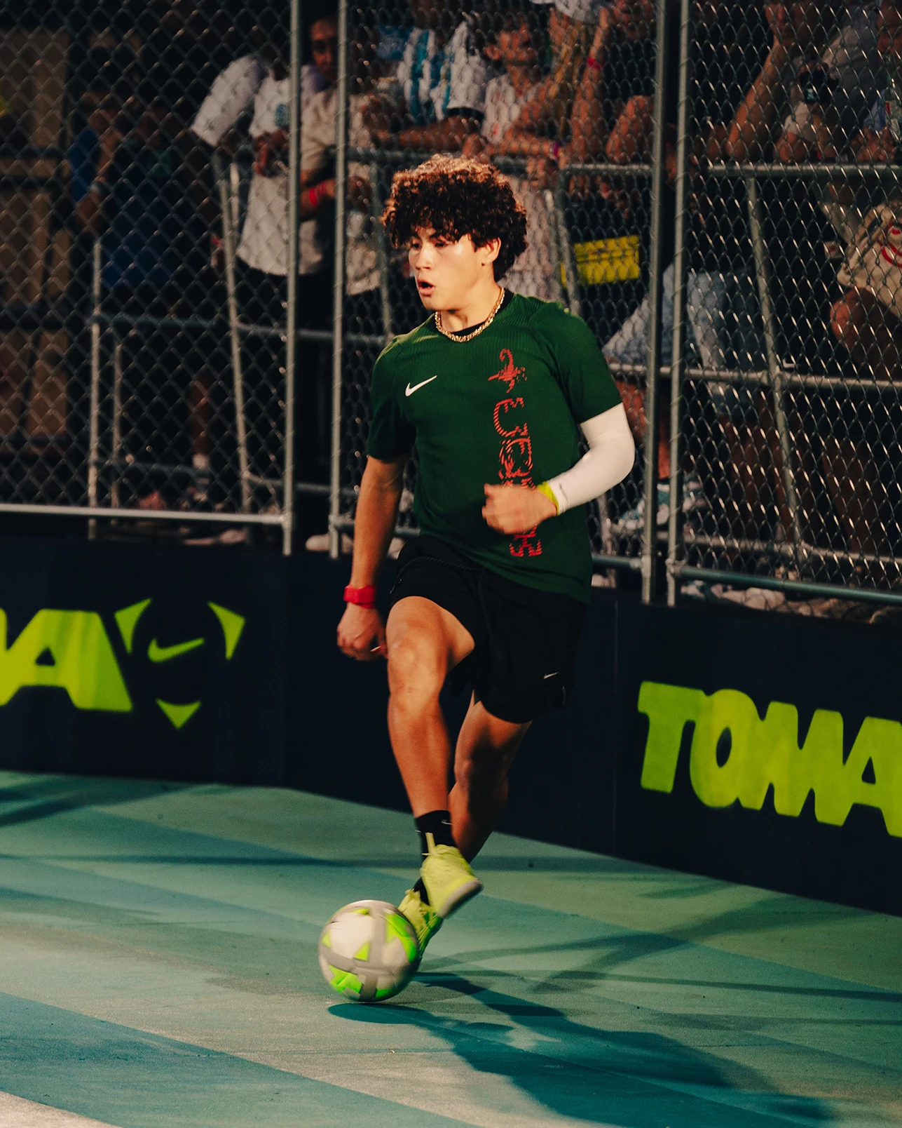 A young soccer player in a green shirt dribbles a ball on an indoor court, with spectators watching behind a chain-link fence.