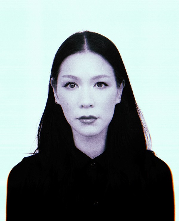 A woman with long dark hair and a neutral expression, wearing a black collared top, faces the camera against a light background.