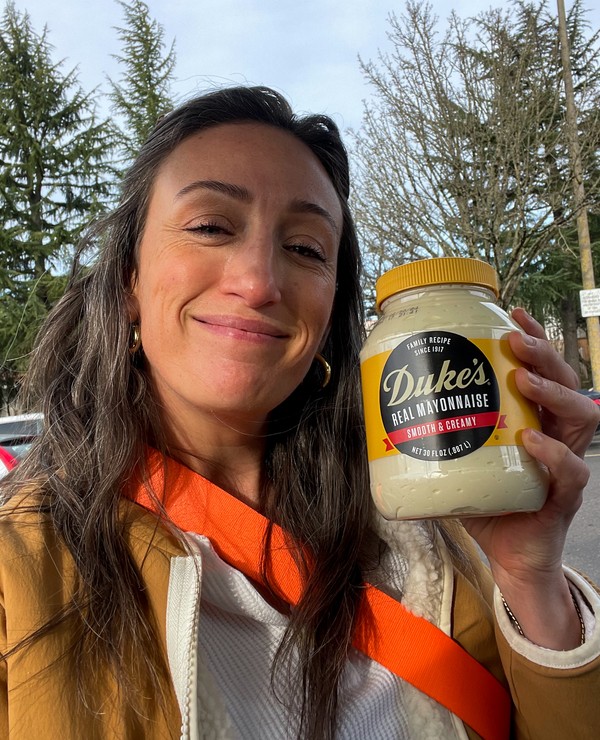 A woman outdoors holds up a jar of Duke’s Real Mayonnaise and smiles at the camera. Trees and a parking lot are visible in the background.