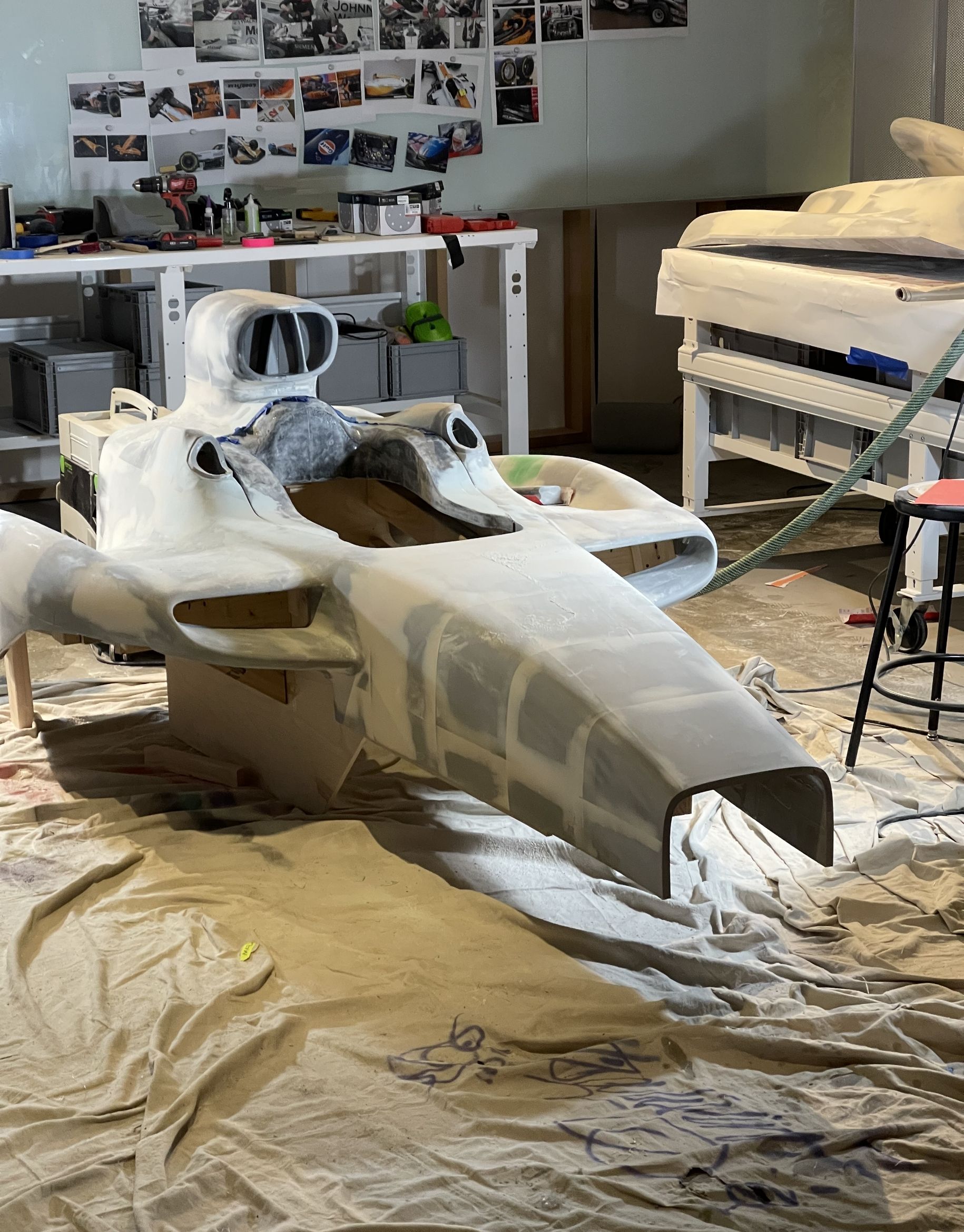 A partially constructed race car body in a workshop, surrounded by tools and photographs.