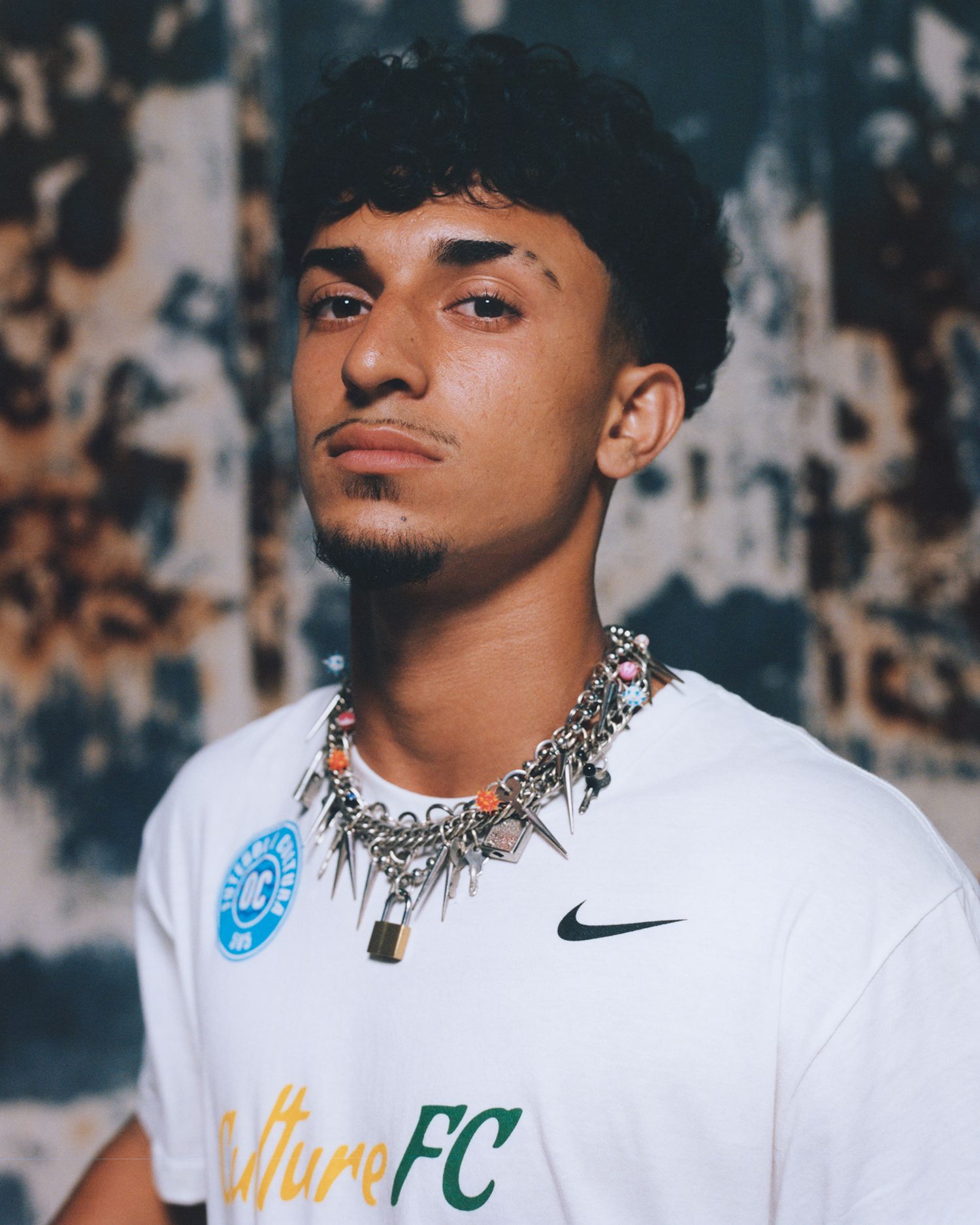 Young man with short curly hair and a thin goatee wearing a white "Culture FC" Nike t-shirt, spiked chain necklaces, and a blue badge, standing in front of a textured wall.