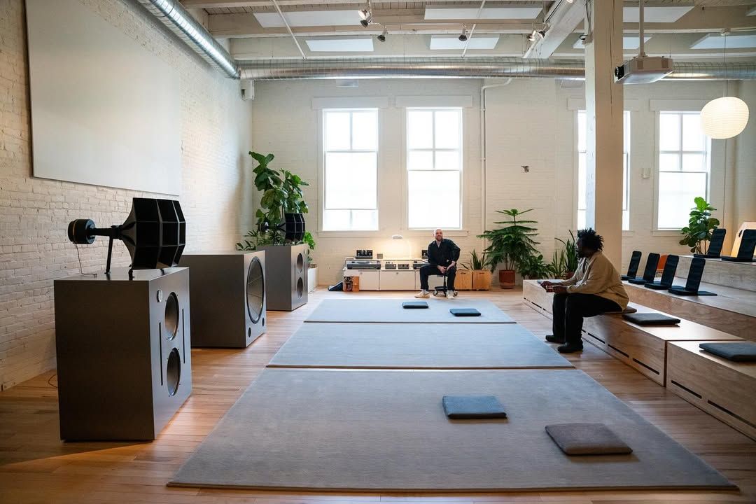 Two people sit in a spacious, minimalist room with large speakers, wooden floors, mats on the floor, and several potted plants, with natural light coming through big windows.