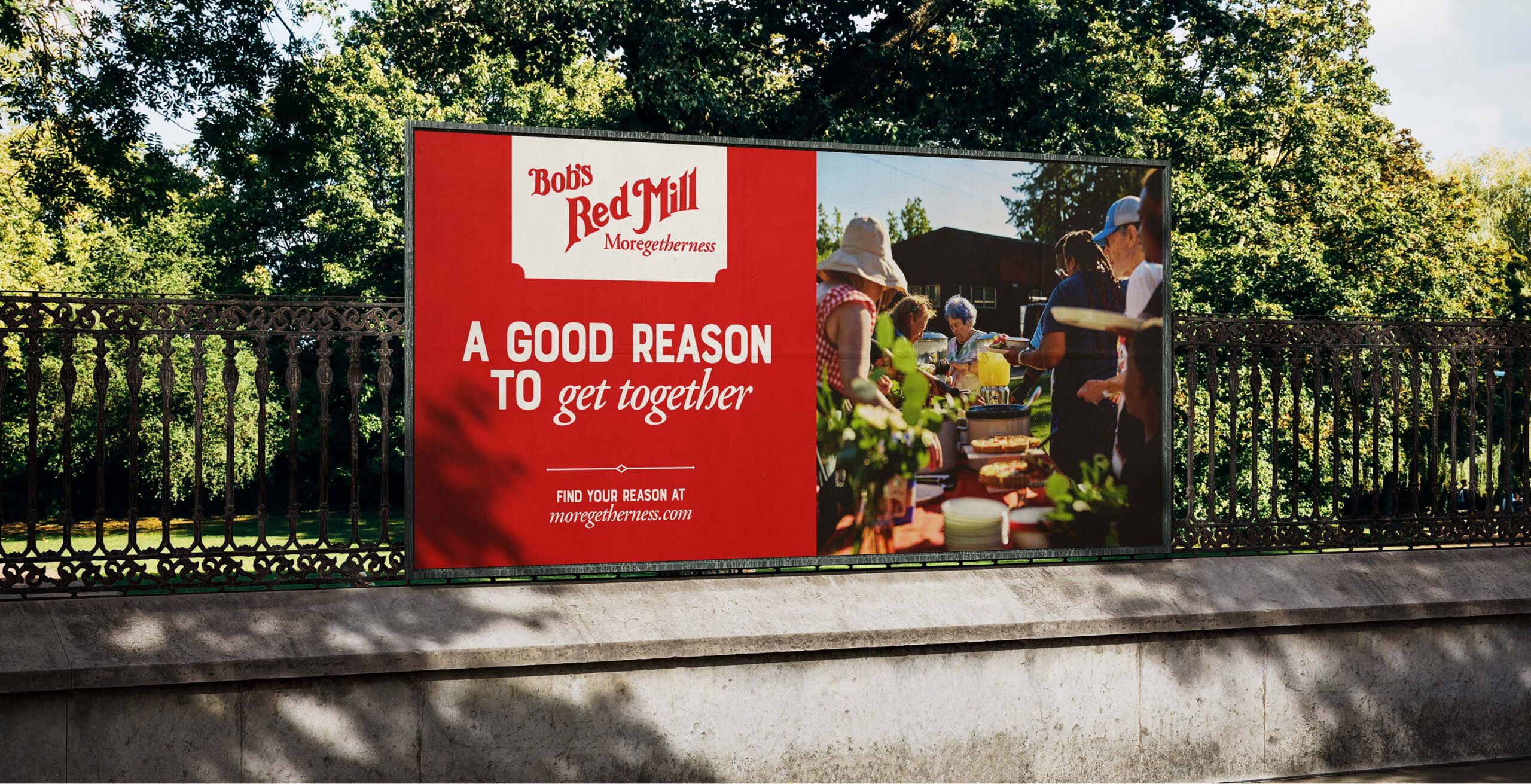 A Bob's Red Mill billboard reads "A Good Reason To Get Together" next to an image of people at an outdoor gathering, set against a backdrop of trees and iron fencing.