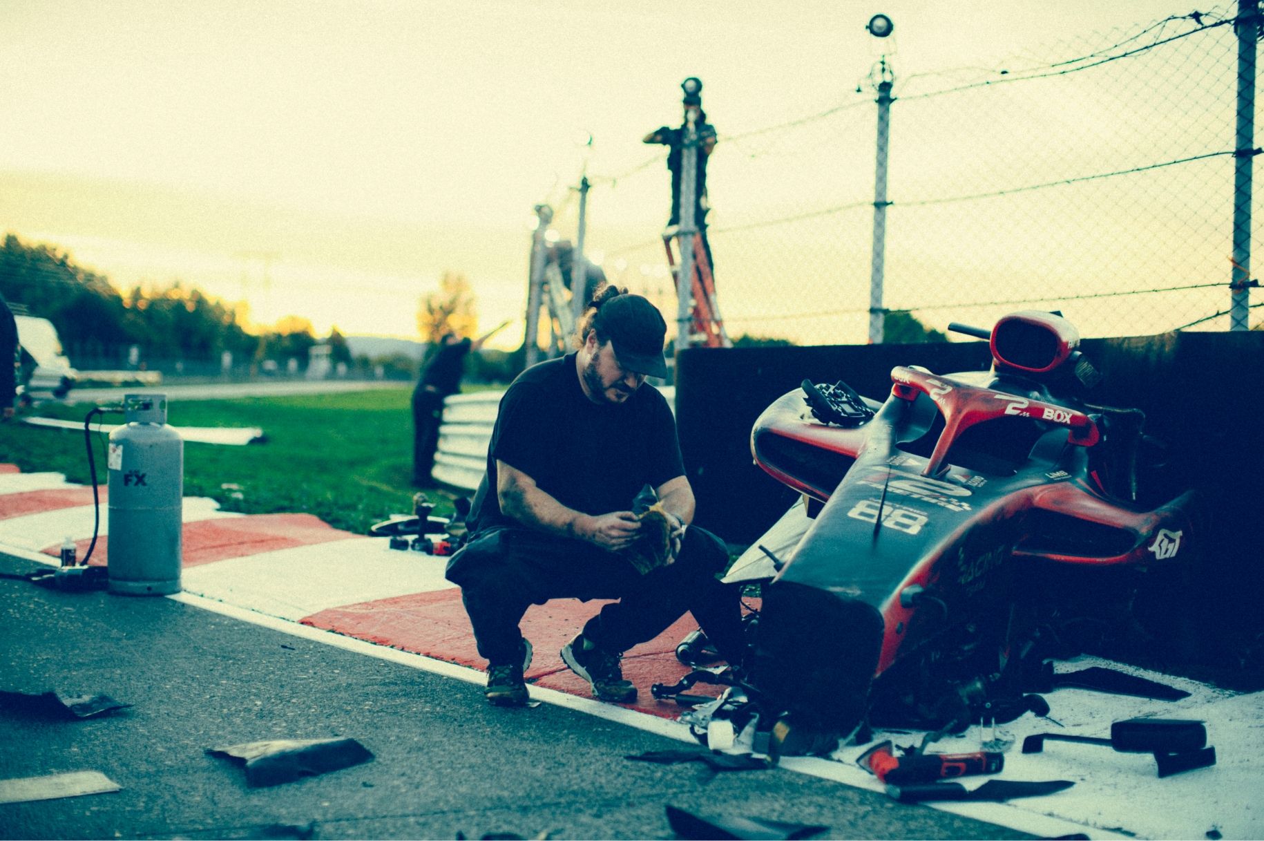 A person examines a damaged race car on the side of a track during sunset. Debris is scattered around.