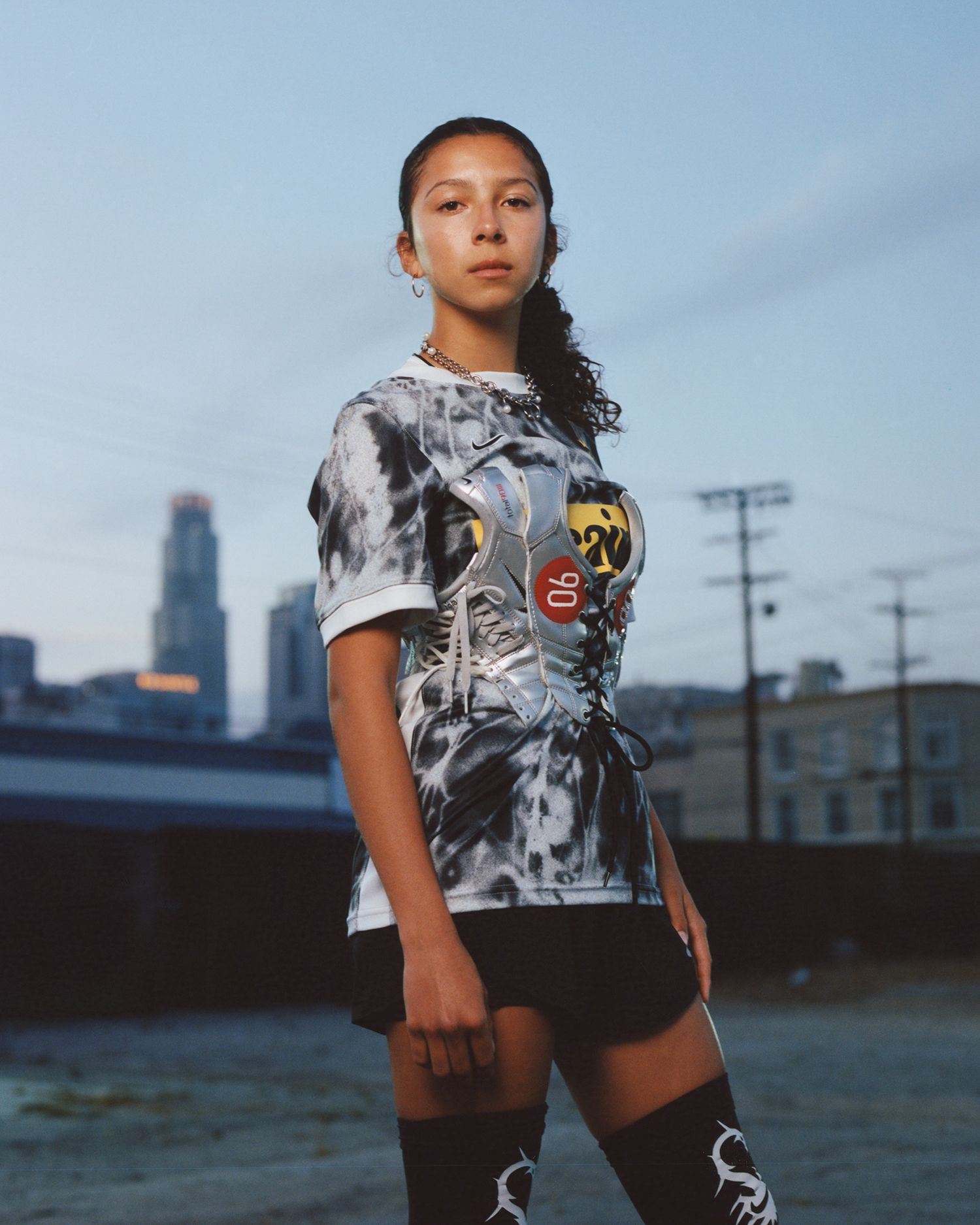 A young woman stands outdoors in front of city buildings, wearing a tie-dye shirt with a graphic design, black shorts, and patterned thigh-high stockings.