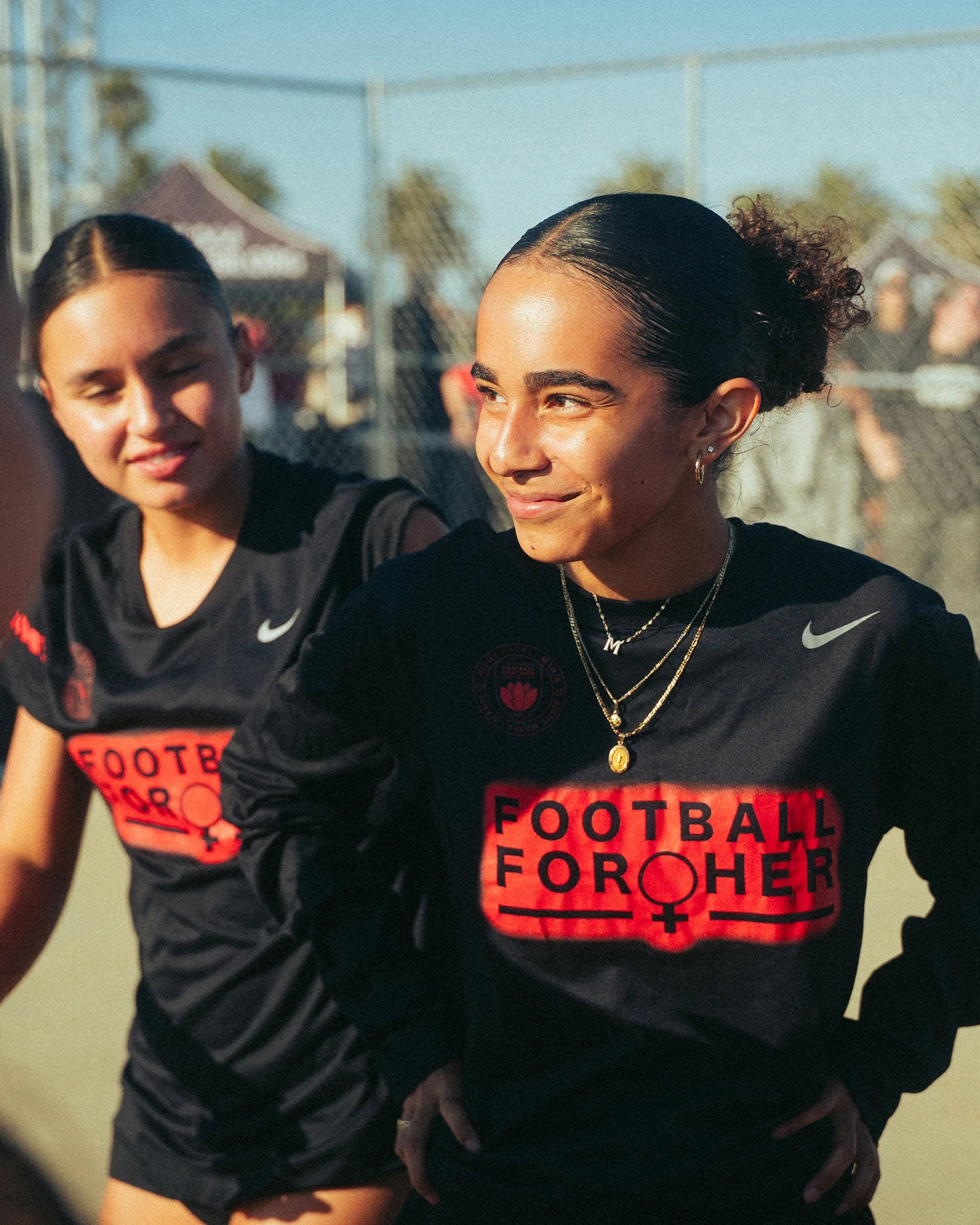 Two young women in black “Football For Her” jerseys stand outdoors on a sunny day, near a chain-link fence, with one smiling and the other looking to the side.