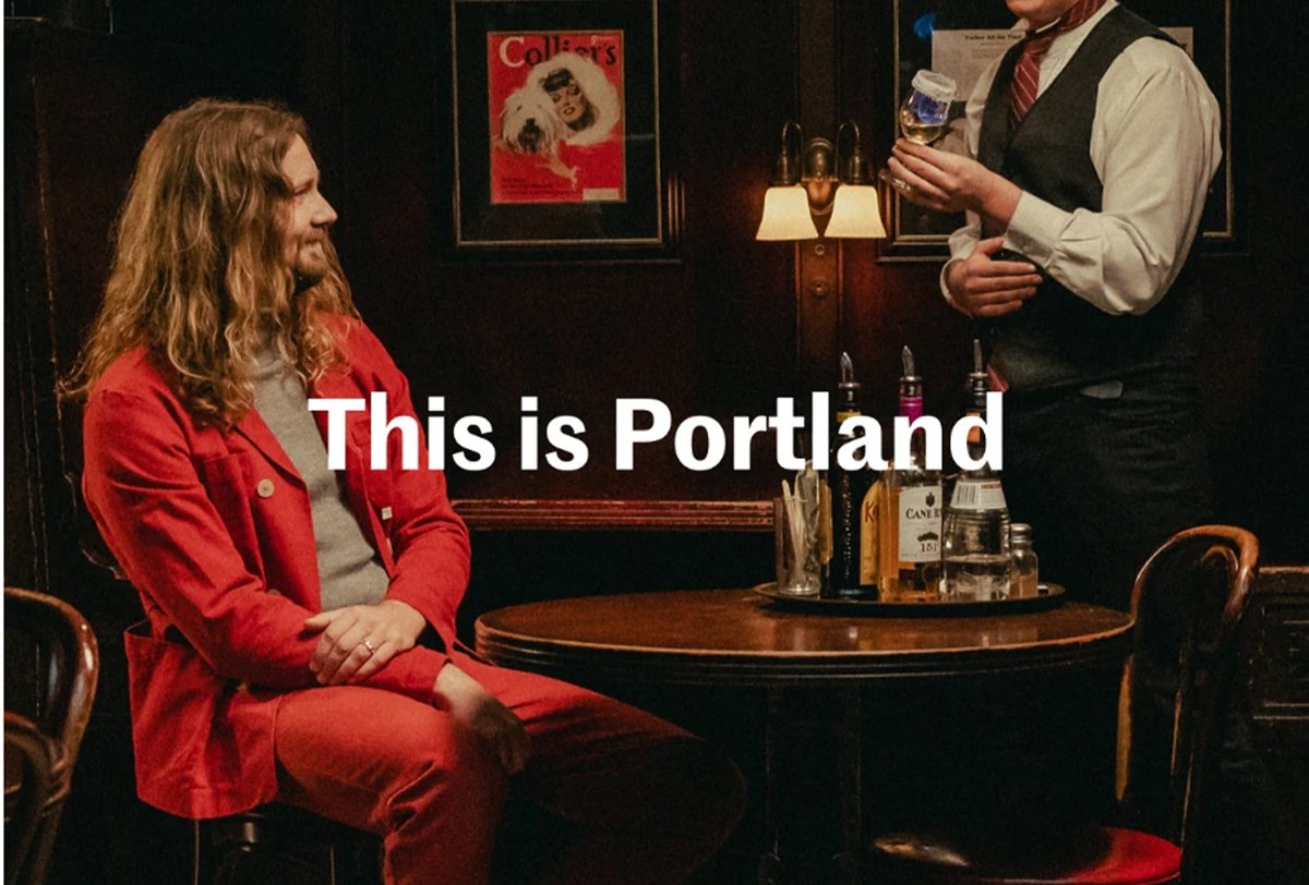 A person in a red suit sits at a table with liquor bottles while a server stands holding a small object; text reads, “This is Portland.”.