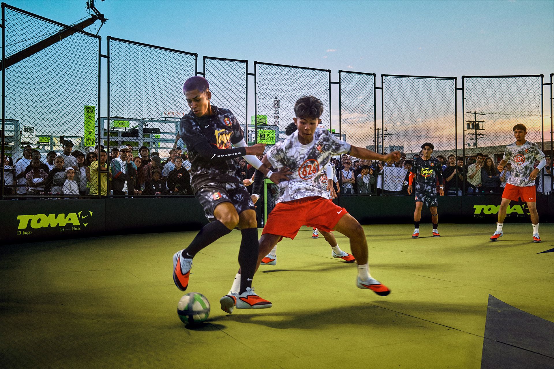 Two soccer players compete for the ball on an outdoor court while a crowd watches behind a fence at sunset. Other players are visible in the background.