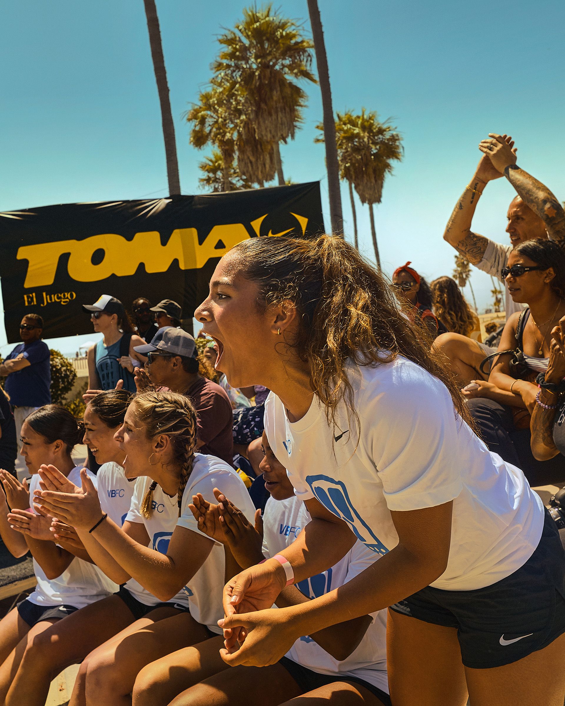 A group of athletes in white uniforms enthusiastically cheer from the sidelines during an outdoor sporting event, with palm trees and spectators visible in the background.