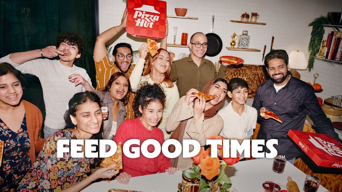 A group of people gather around a table, smiling and eating Pizza Hut pizza. Pizza boxes and food are on the table. Text reads, “FEED GOOD TIMES.”.