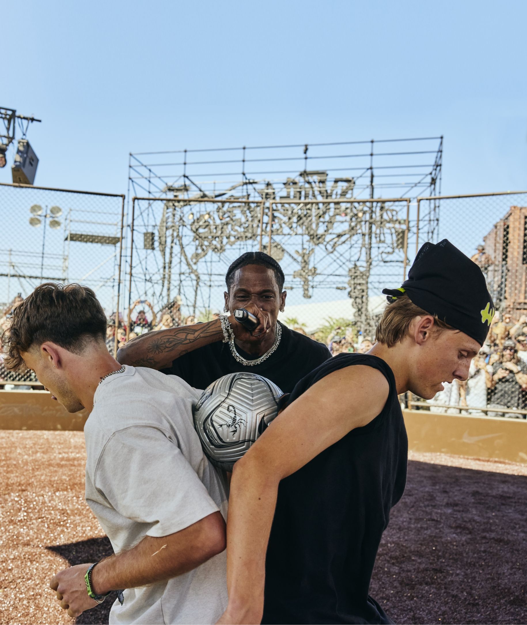 Three people interact at an outdoor event; one holds a microphone, while two press a soccer ball between their backs. A crowd and metal framework are visible in the background.