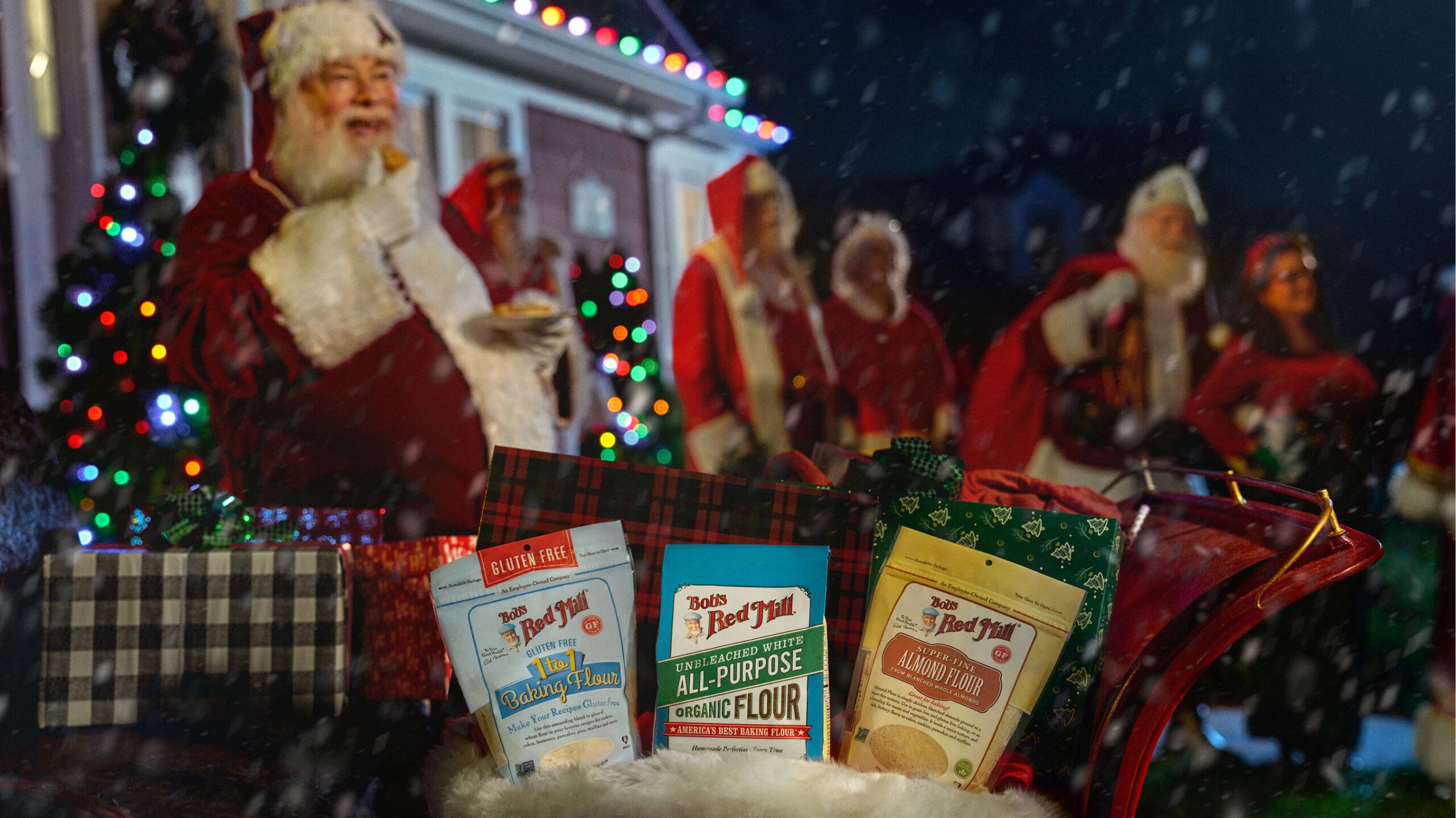 Several bags of Bob’s Red Mill flour are displayed in a festive basket with wrapped gifts, while people dressed as Santa Claus stand in the background near a decorated house at night.
