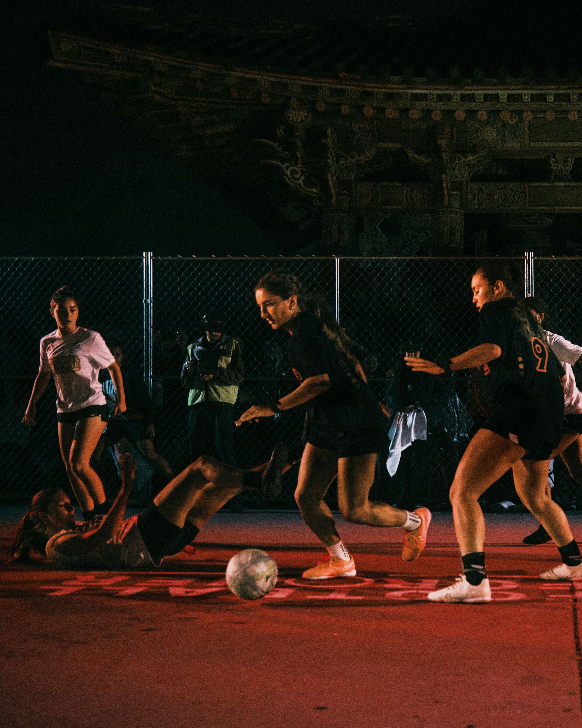 A group of women play soccer at night on a red court, with one player on the ground and others competing for the ball. A fence and traditional building are visible in the background.