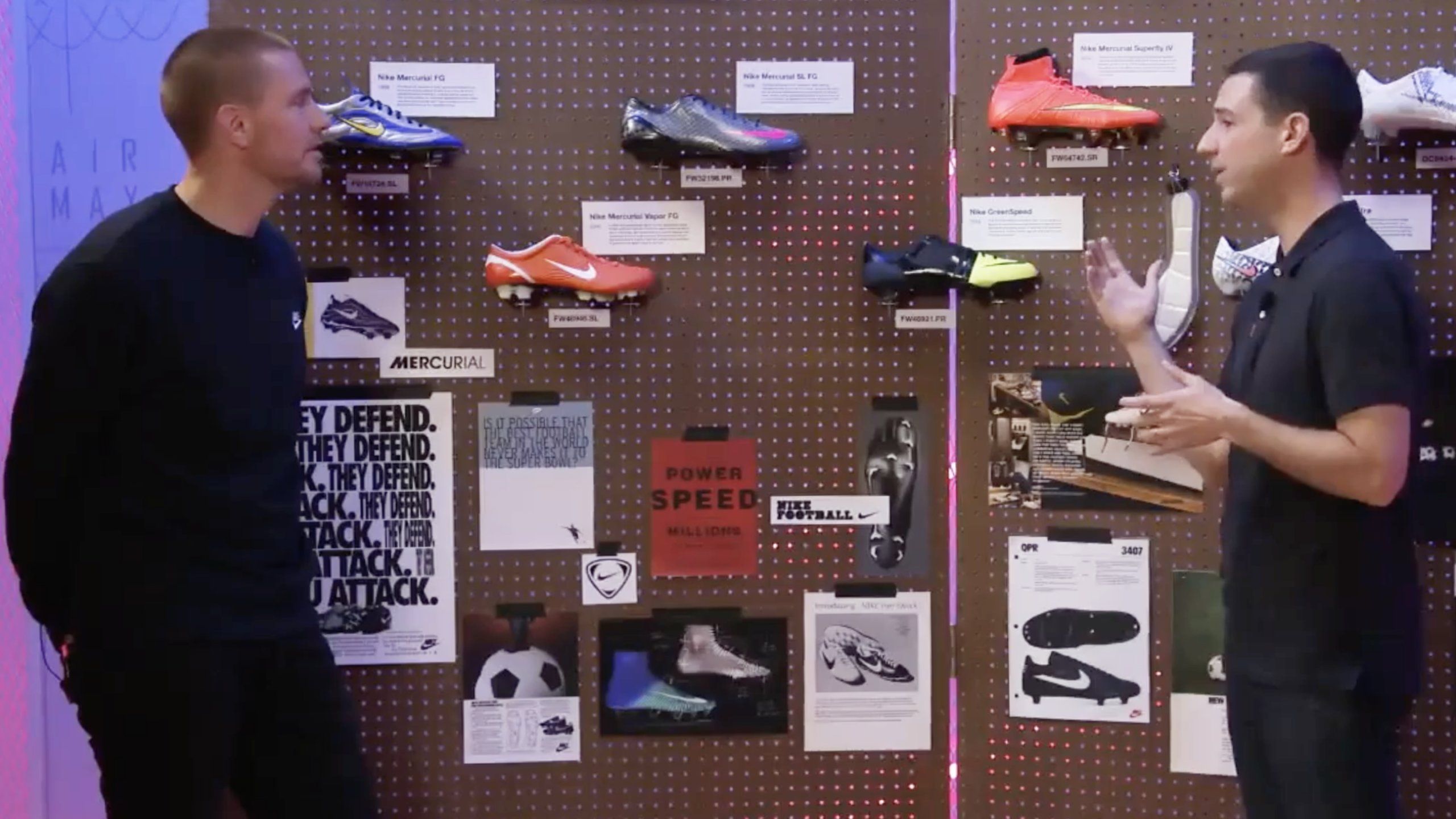 2 Event host speaking to each other at pegboard of with past promotional ads and football boots that Nike has made through the years