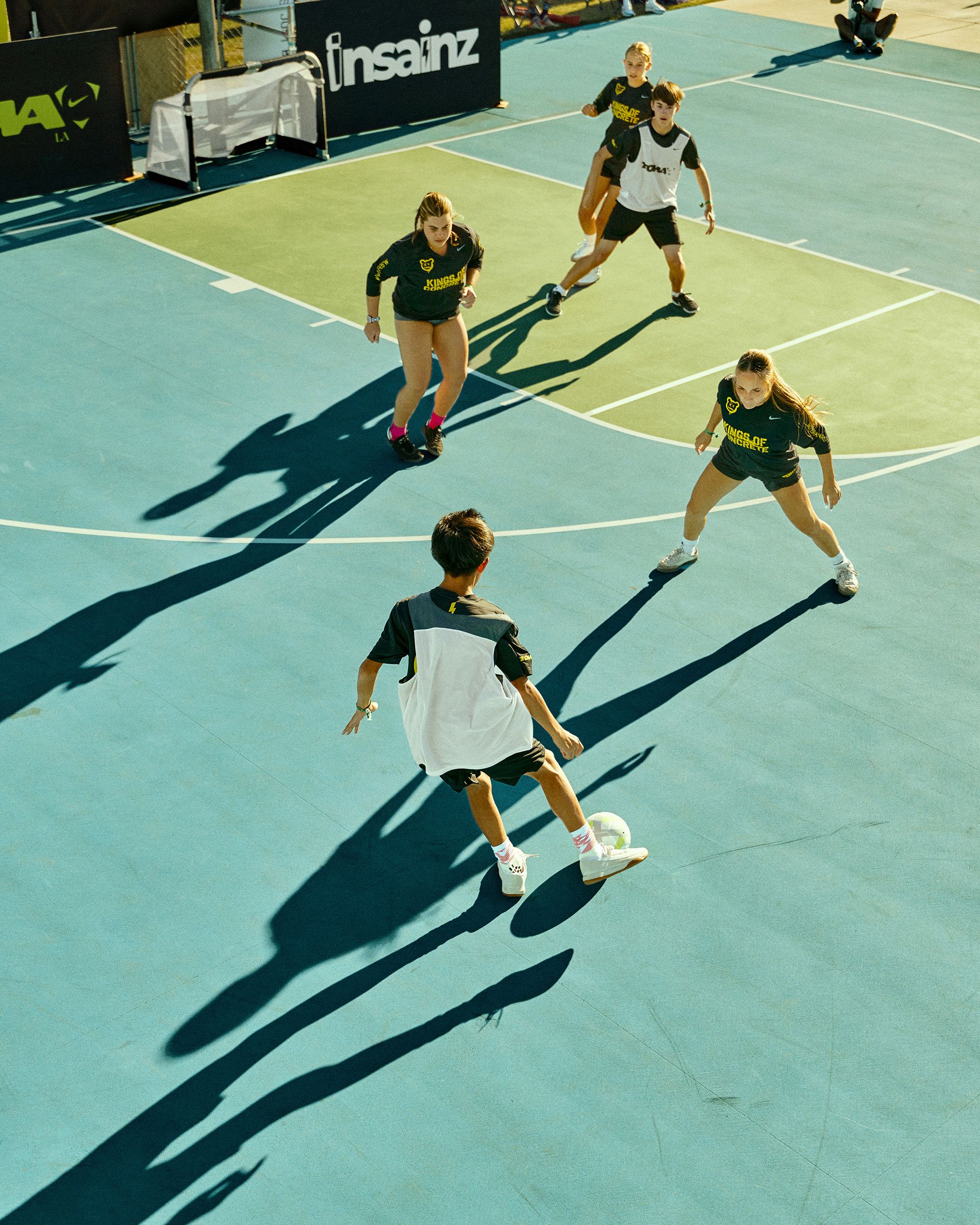 A group of young people play soccer on an outdoor blue court, with one player dribbling the ball toward four defenders.