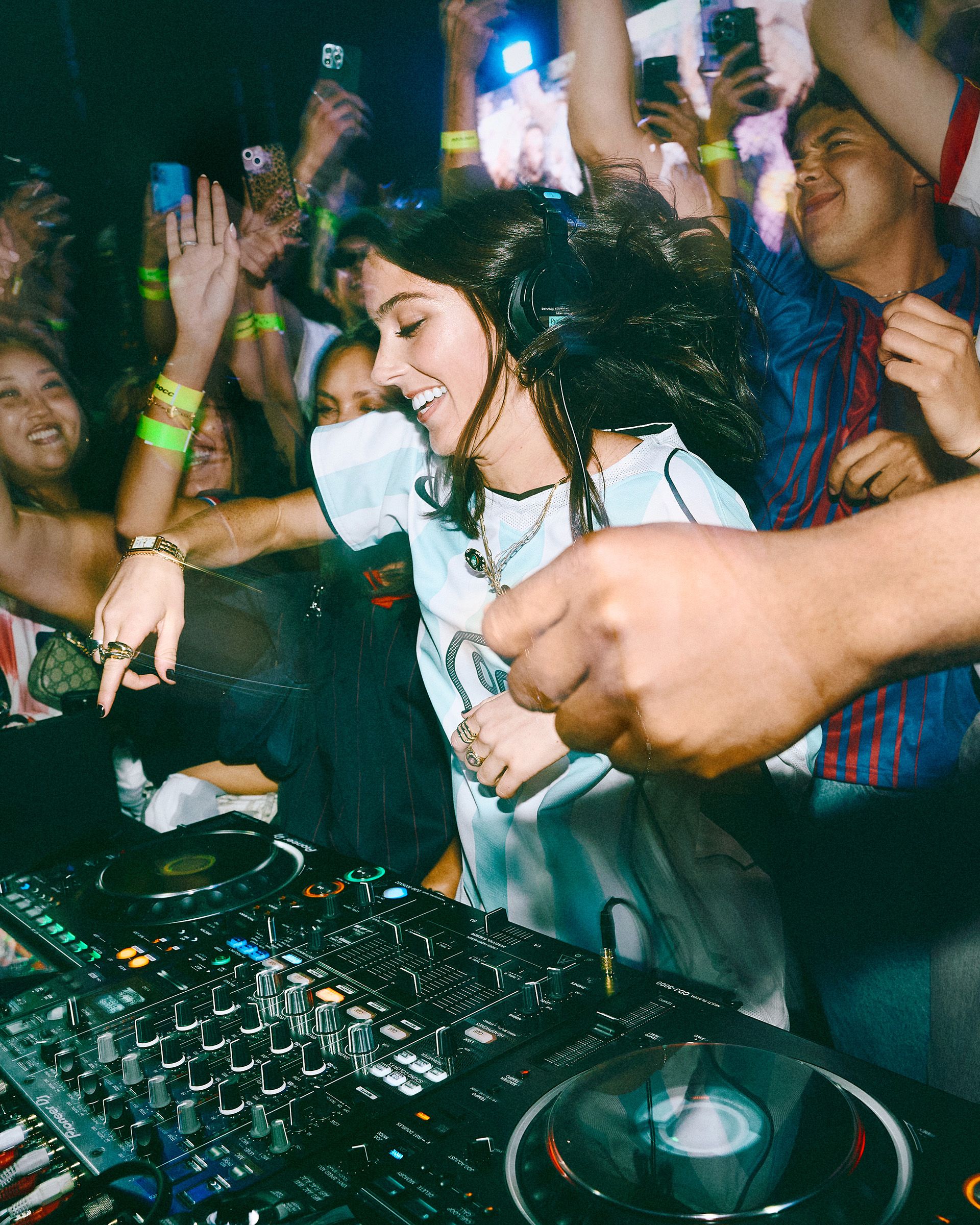 A DJ wearing headphones plays music at a crowded party, with people cheering and raising their hands around the DJ booth.
