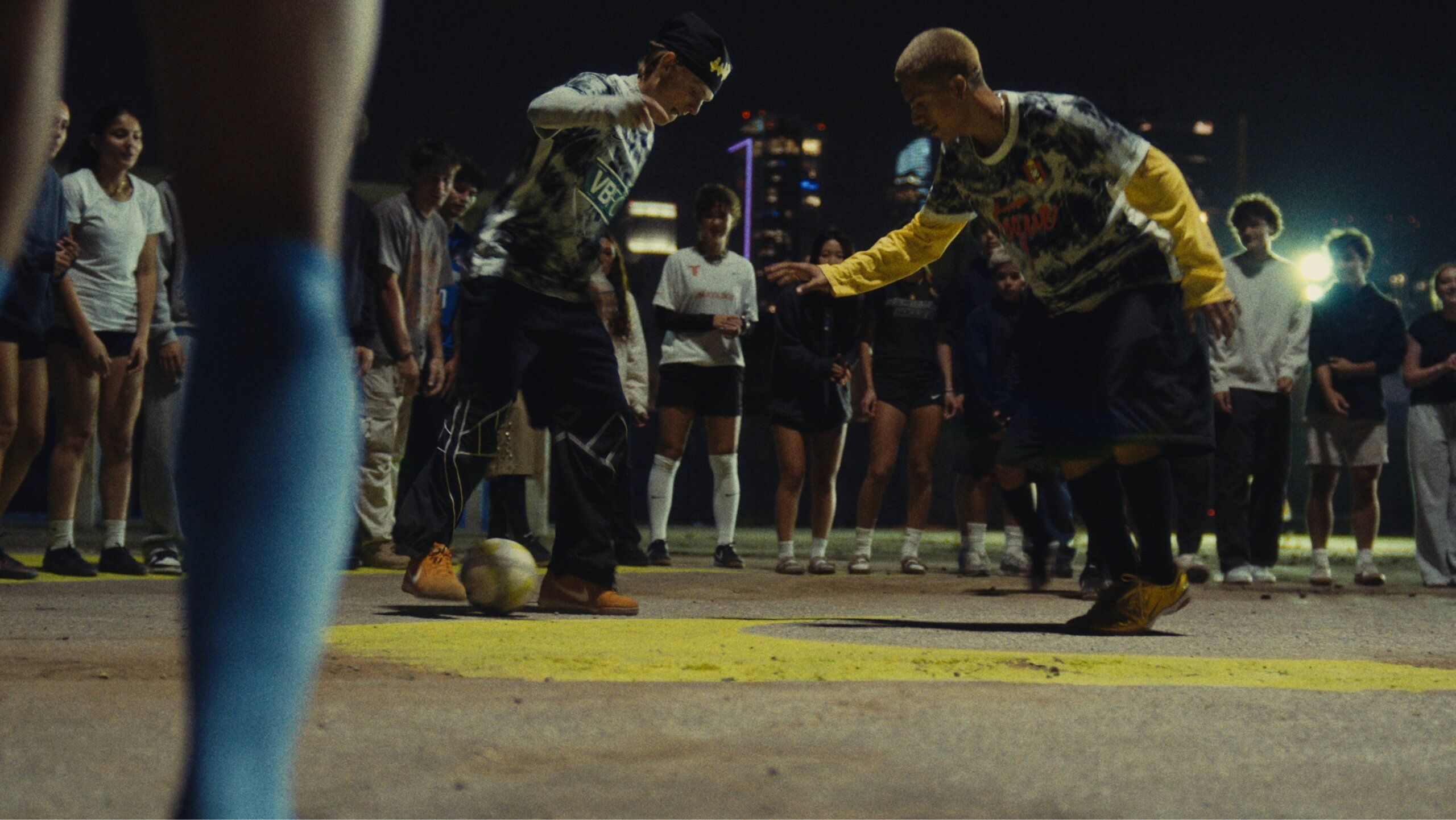Two people play soccer on an outdoor court at night, surrounded by a group of onlookers; city lights are visible in the background.