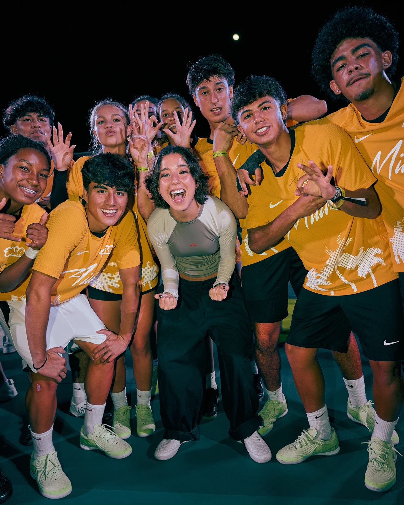 A group of young people in yellow sports shirts pose together and smile for the camera on an outdoor court at night.