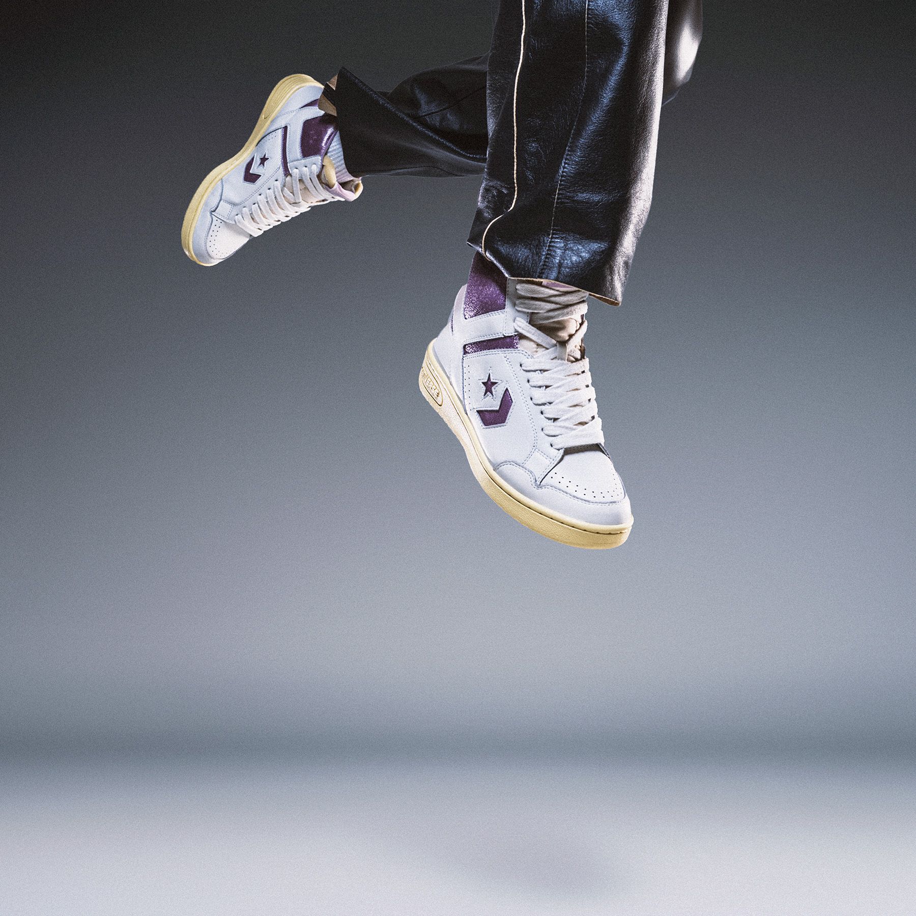 Person wearing white and purple high-top Converse sneakers with yellow soles, captured mid-jump against a gray background.