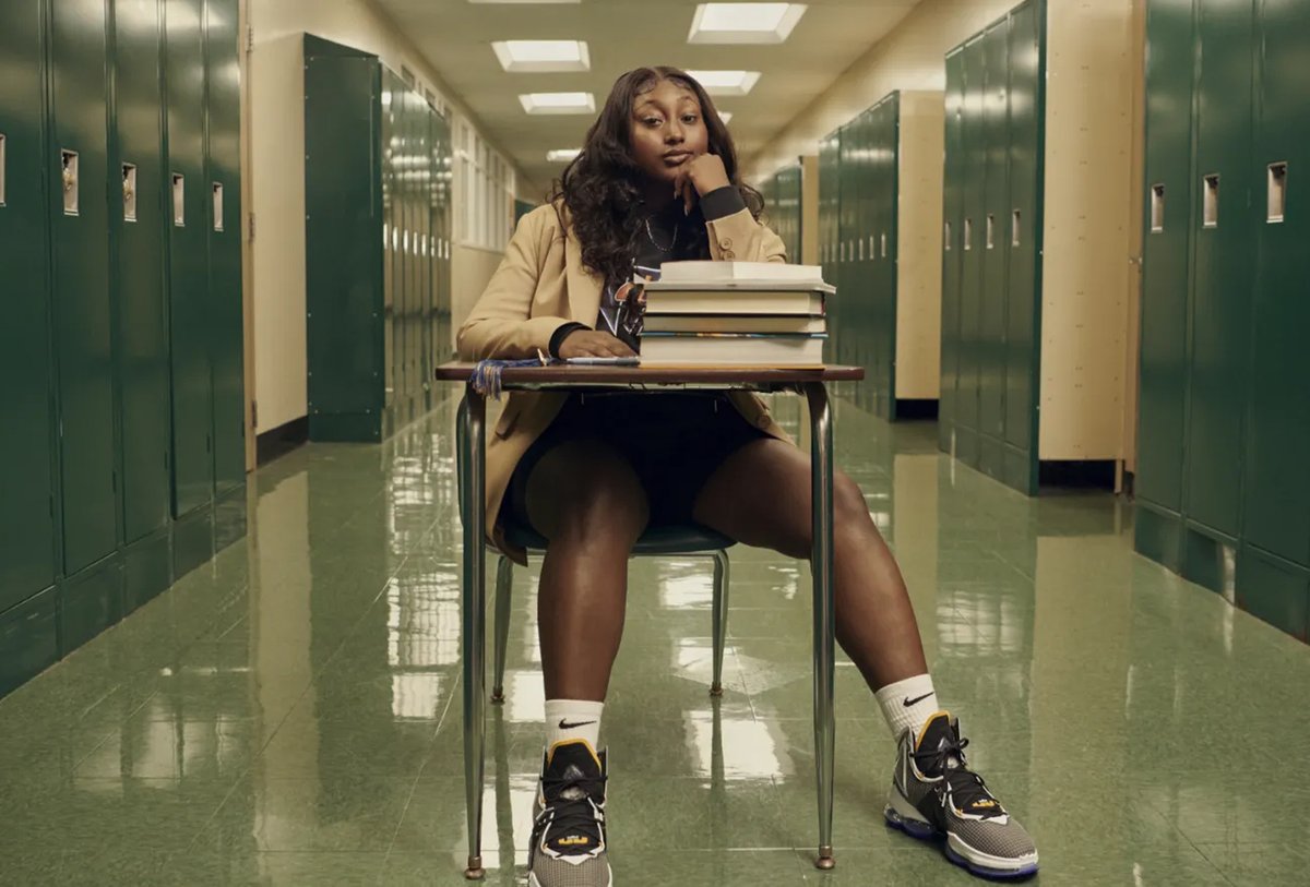 A student sits at a desk with stacked books in a school hallway, resting her head on one hand and looking forward.