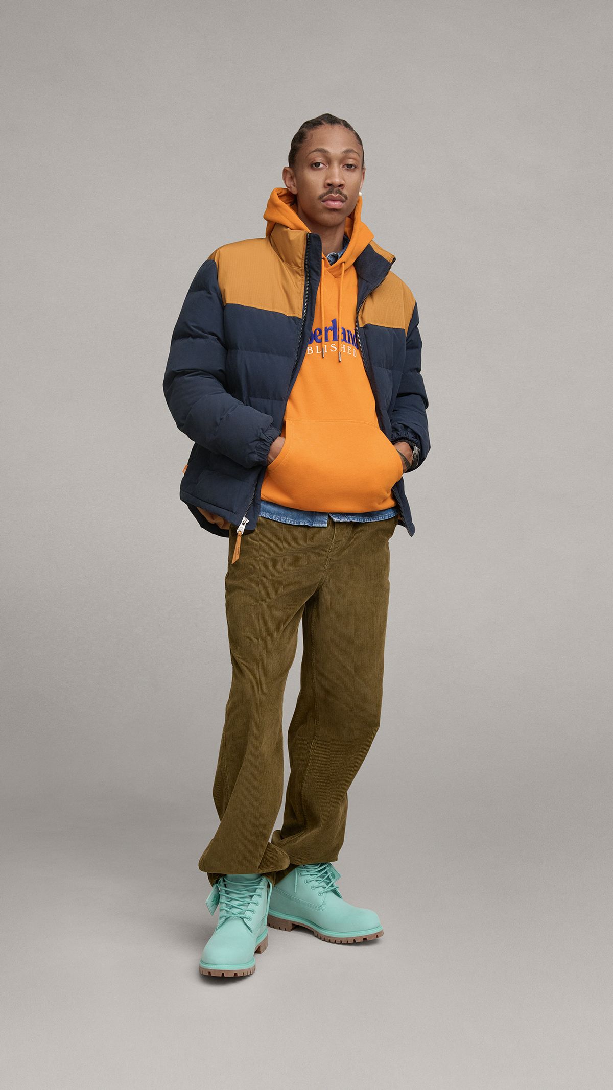 Man standing with Timberland orange and navy blue winter jacket over orange hooded sweatshirt, brown pants and Holiday light blue color way Timberland boots