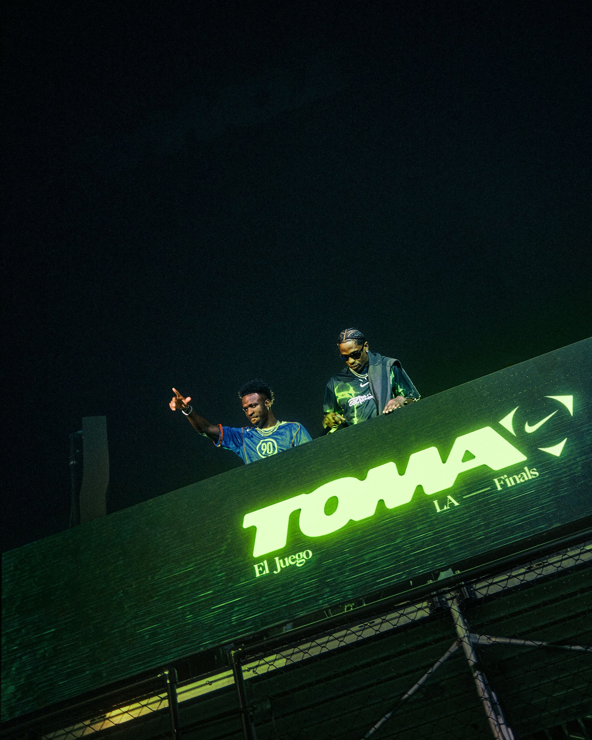 Two men stand behind a large illuminated sign that reads "TOMA El Juego LA Finals" at an elevated venue, with one pointing into the crowd.