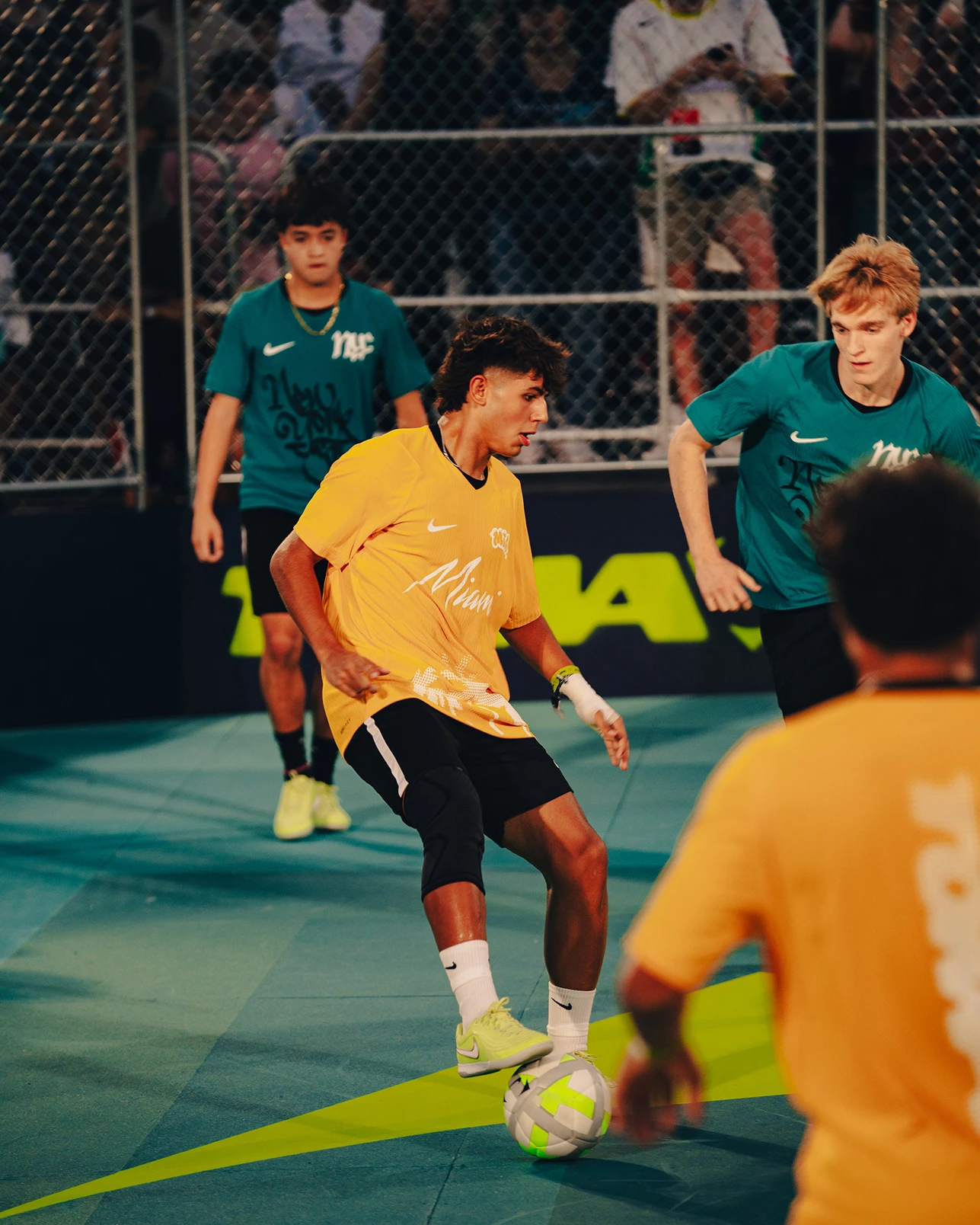 Four young men play indoor soccer on a blue court, with two in yellow jerseys and two in teal jerseys, as the player in yellow prepares to kick the ball.