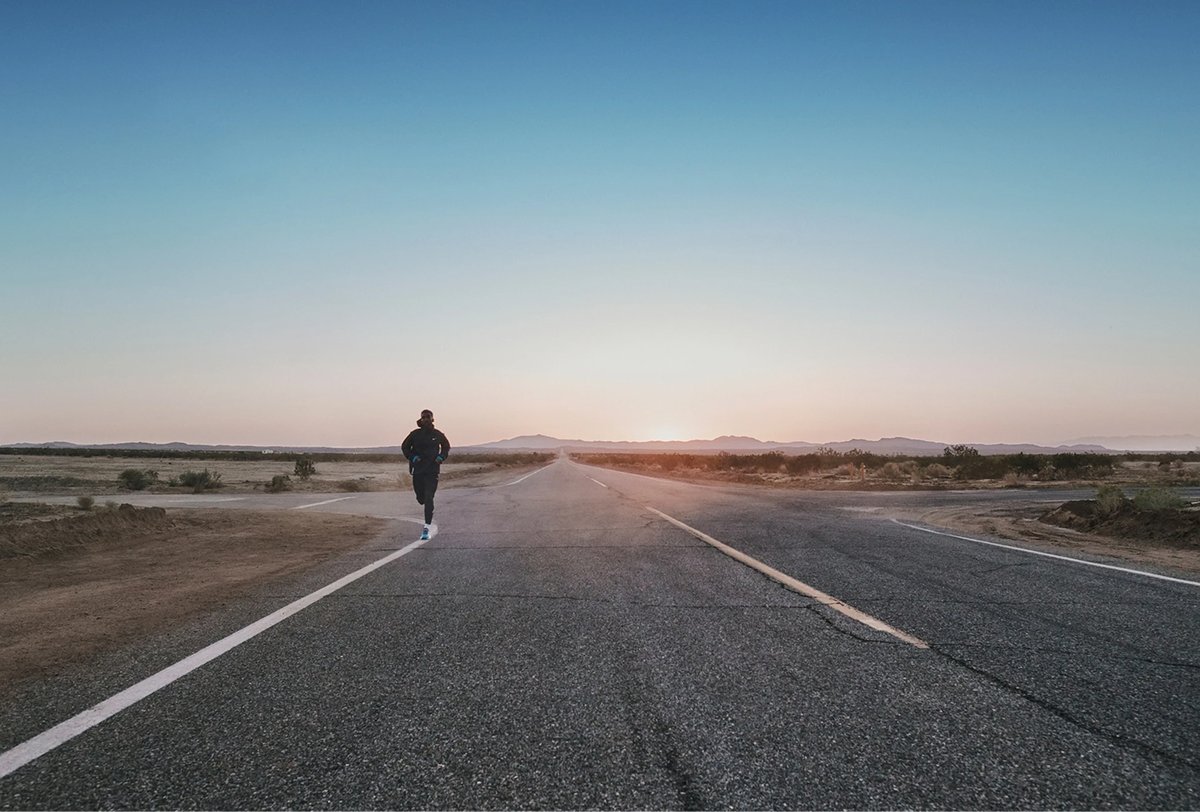 A person runs alone on a deserted road through a dry landscape at sunrise or sunset, with clear sky and distant mountains in the background.
