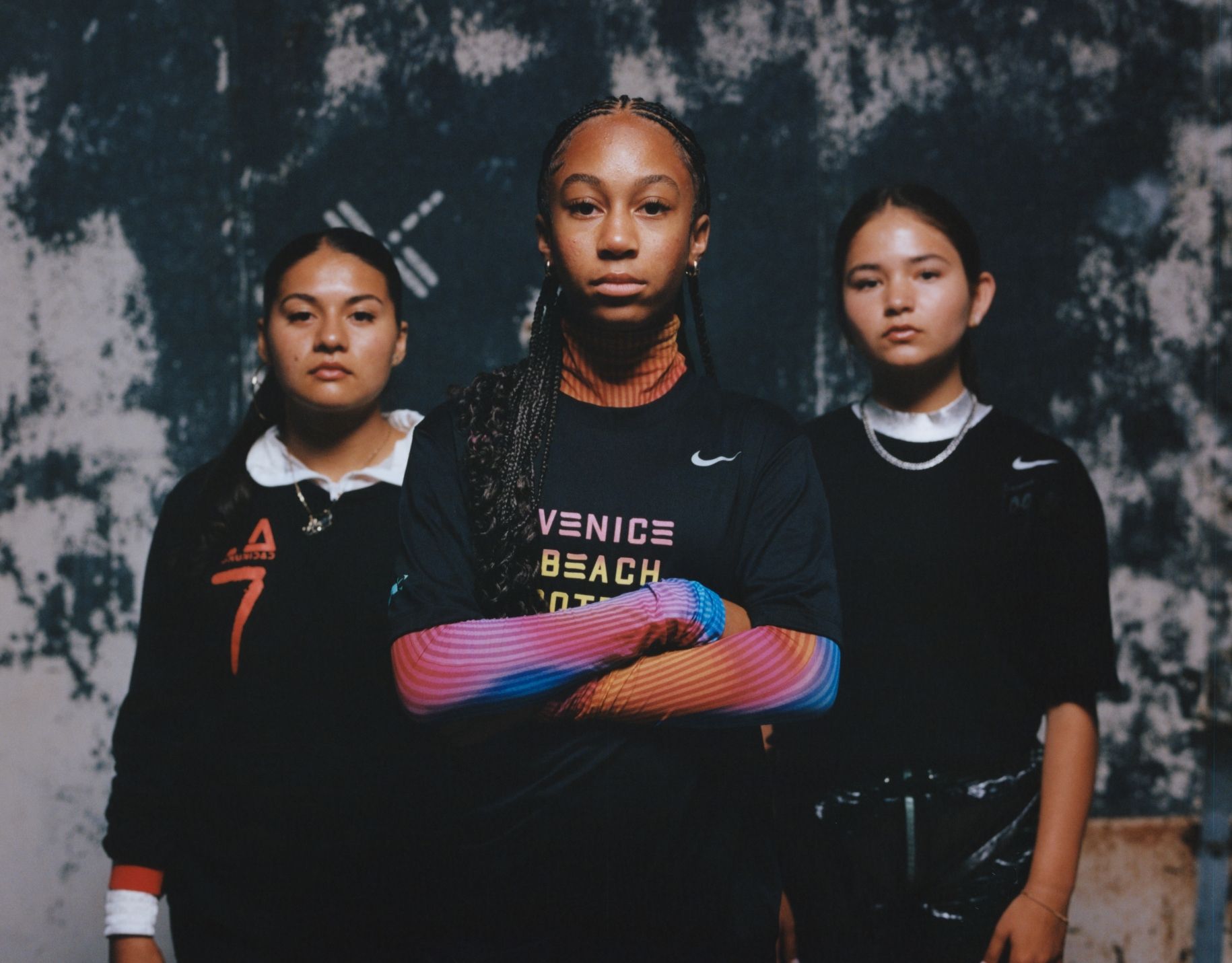 Three young women stand side by side against a dark, mottled background, facing the camera with serious expressions. The central figure has crossed arms and wears a "Venice Beach" shirt.