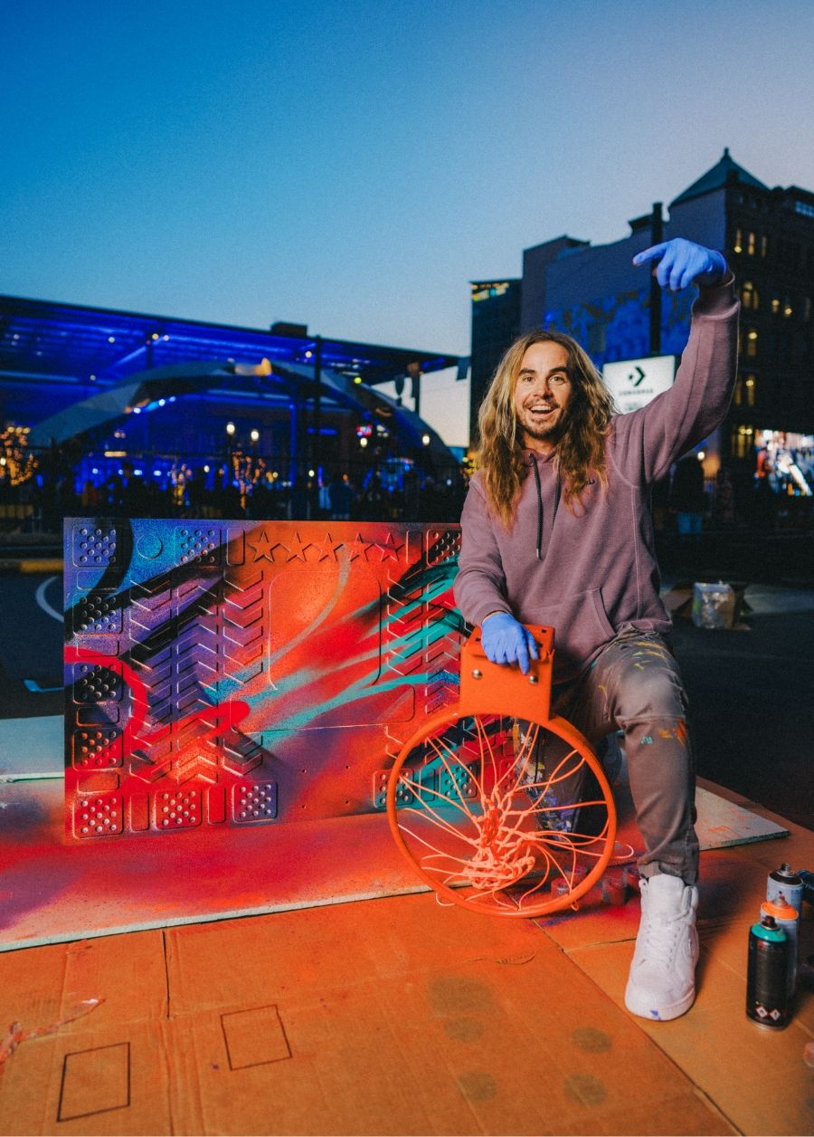 Aritst, Madsteez, kneels with his custom spray painted basketball backboard and basketball hoop