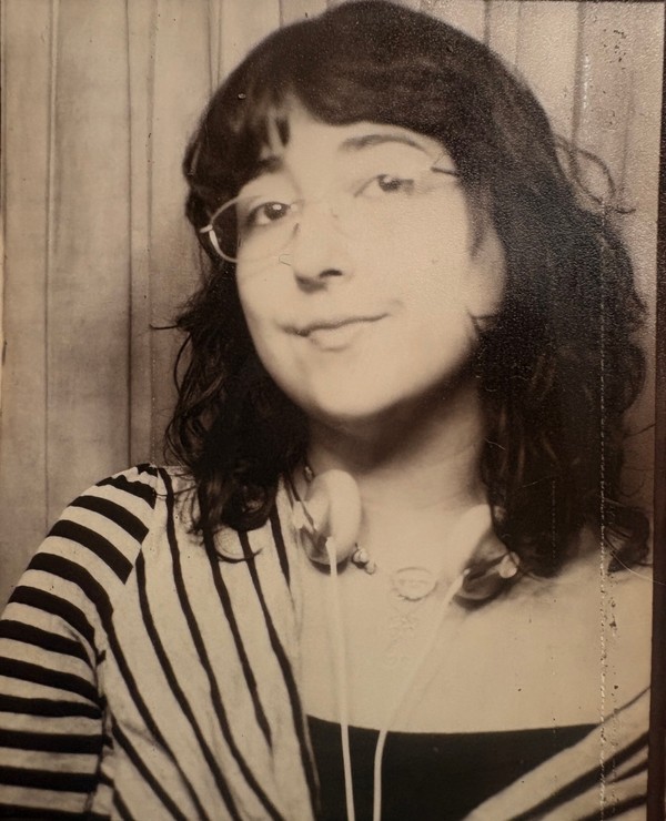 A person with glasses and wavy hair wears headphones around their neck and a striped top, standing in front of a plain backdrop in a black and white photograph.