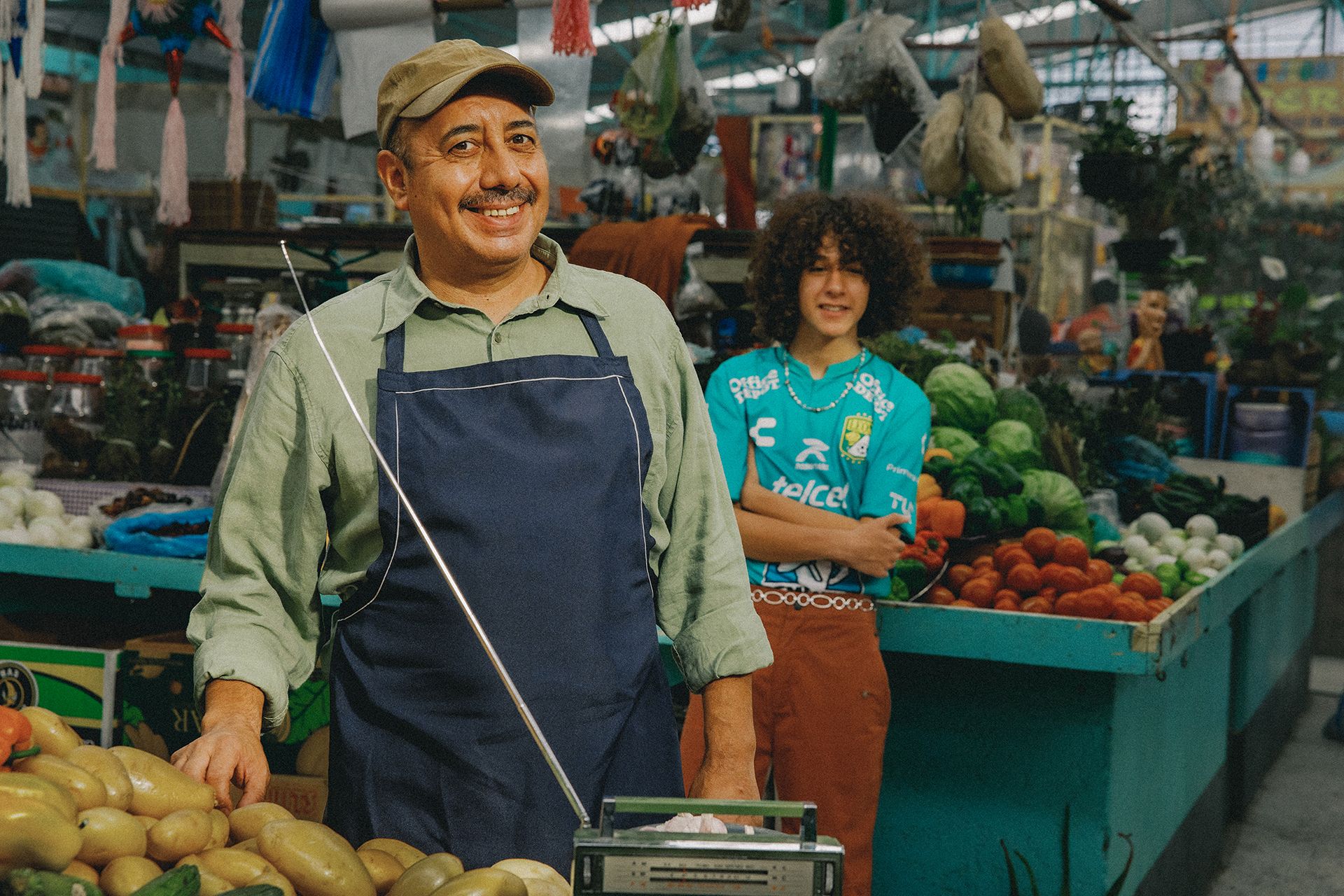 A vendor in a blue apron and cap smiles behind a stall of root vegetables in an indoor market. Another person stands in a blue shirt with arms crossed, amidst various produce.