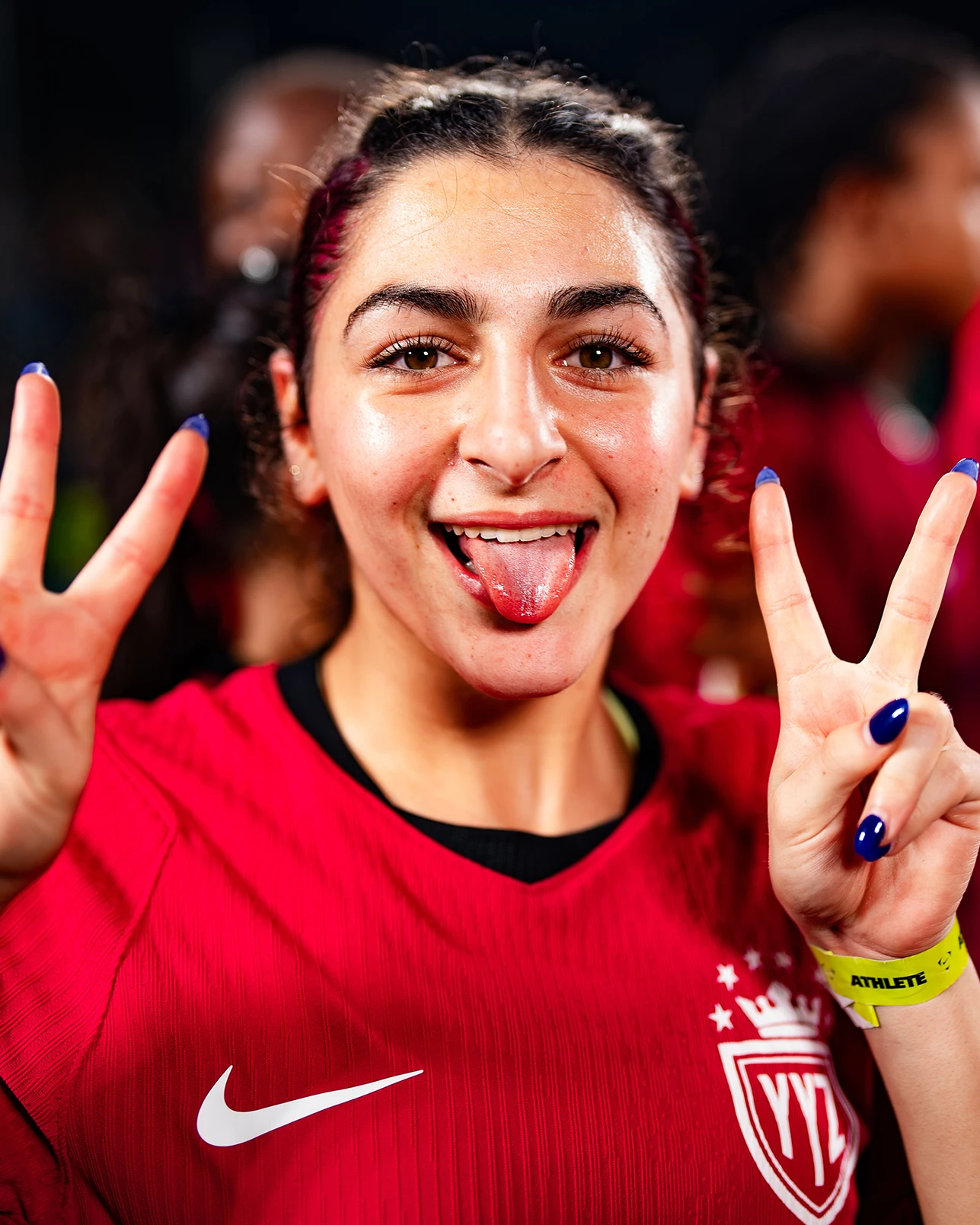 A soccer player in a red jersey holds up two peace signs and sticks out her tongue, with others blurred in the background.