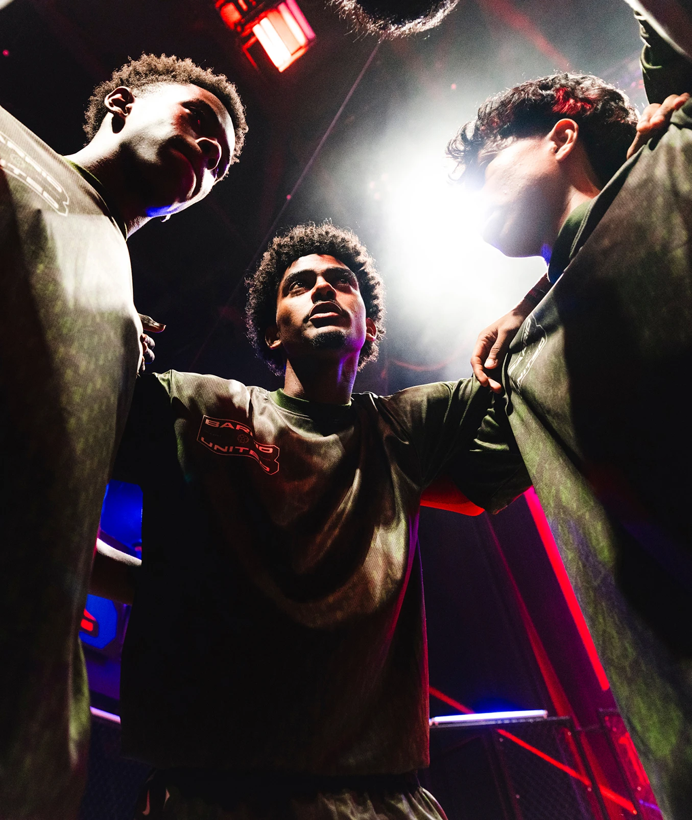 Three people in sports uniforms stand in a huddle under bright lights, arms on each other’s shoulders, with dramatic lighting and a dark background.