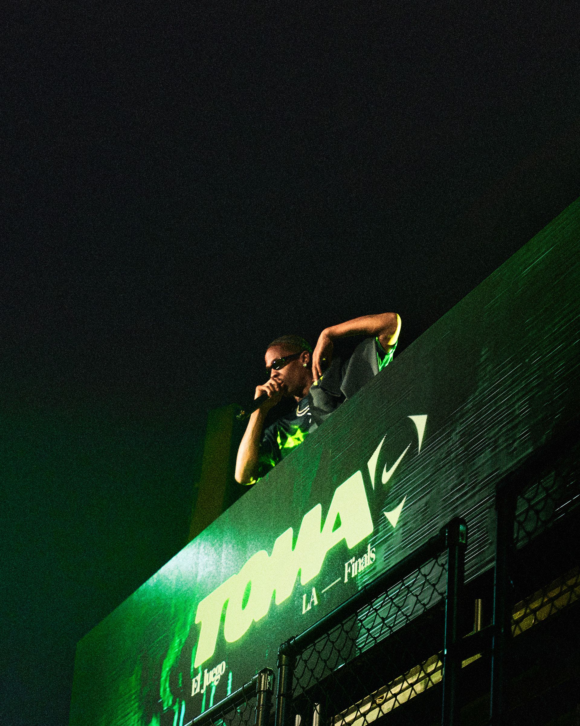 Travis Scott wearing sunglasses leans over a railing at an event, illuminated by green light. The railing features the word "TOMA" and other event branding.