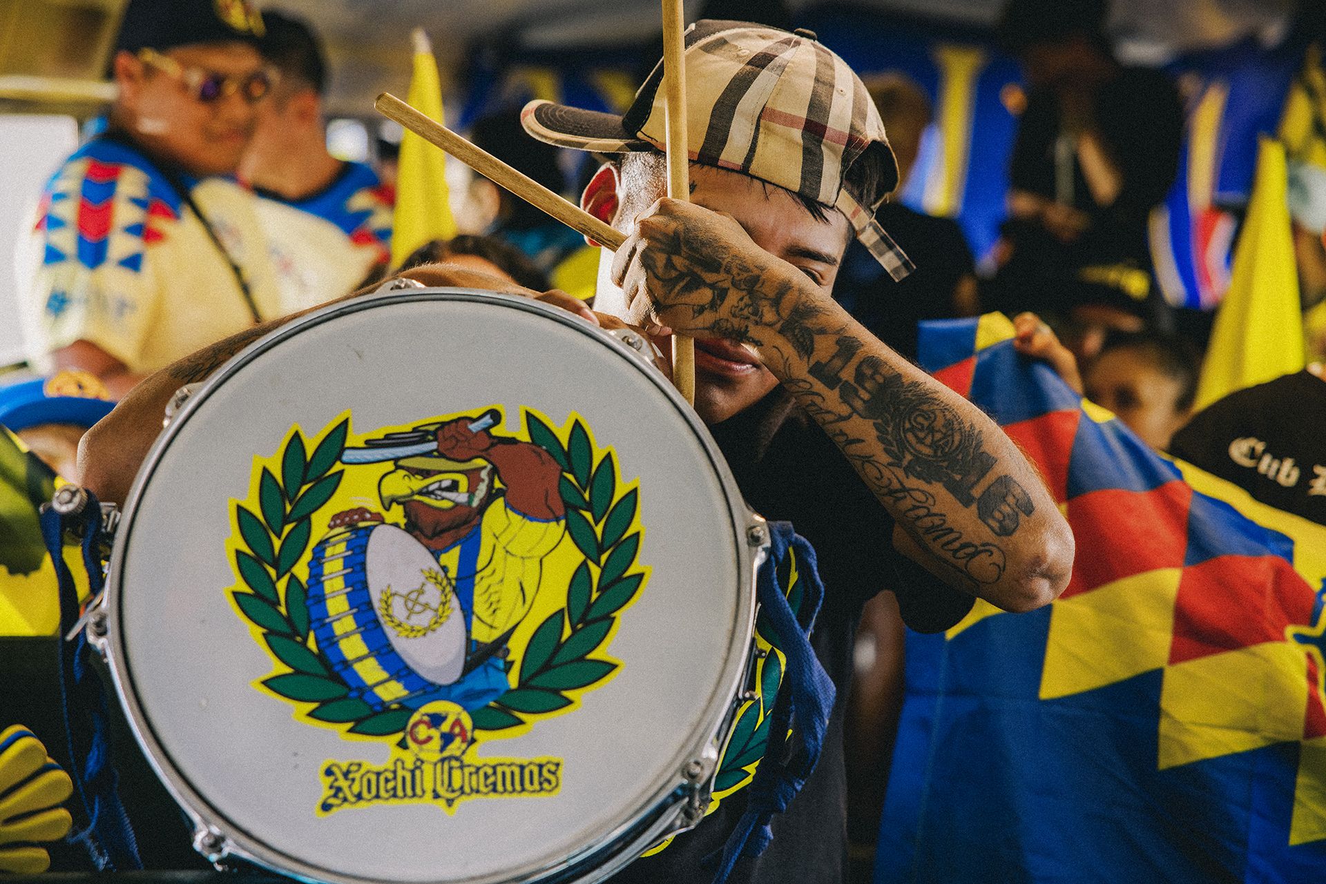 A person with tattoos plays a large drum adorned with a colorful emblem, surrounded by people holding flags and wearing coordinated clothing, possibly during a lively event or celebration.